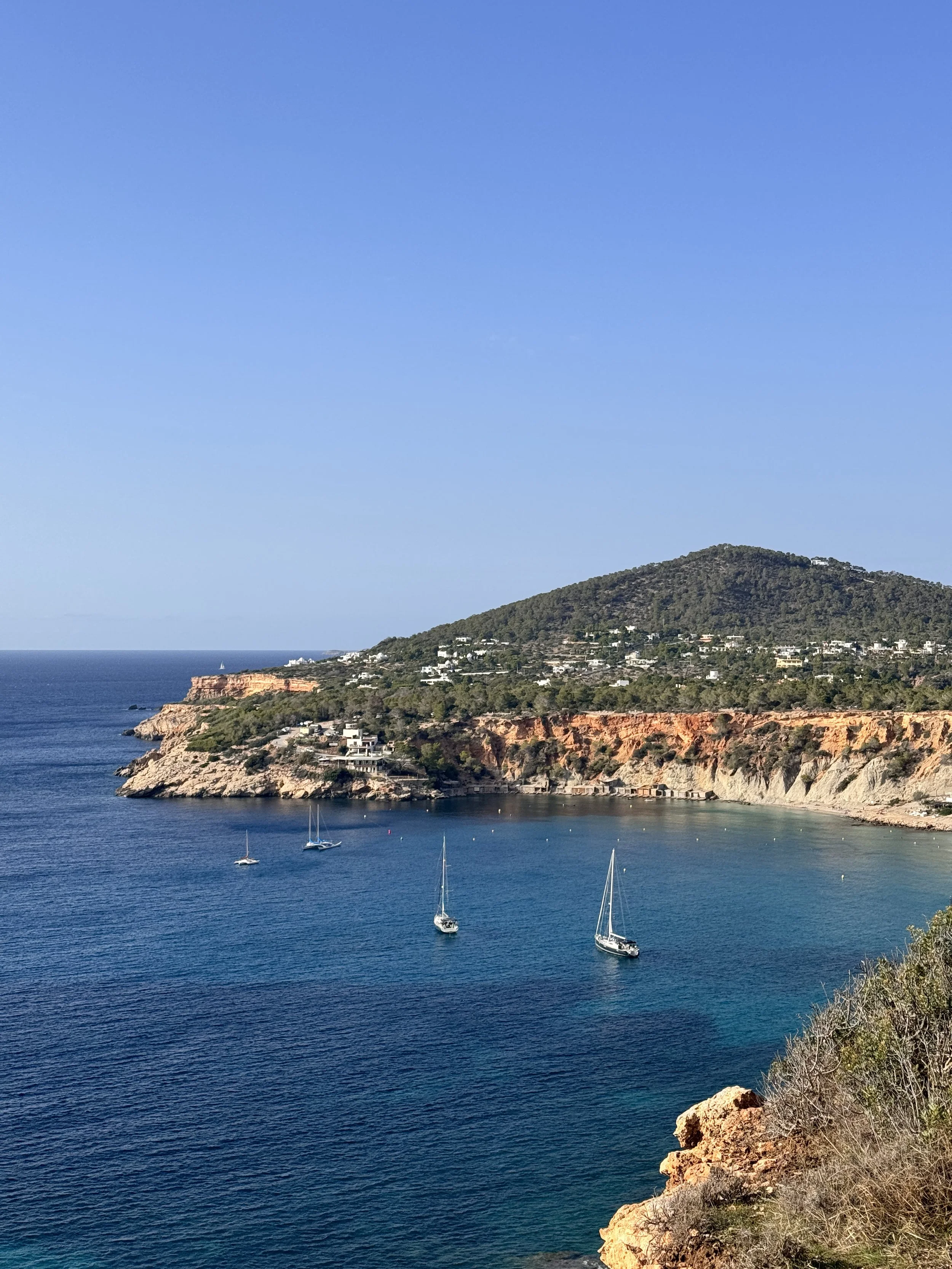 A view of a coastal bay with three sailboats on calm blue water, rocky cliffs, and hillside with houses under a clear blue sky.