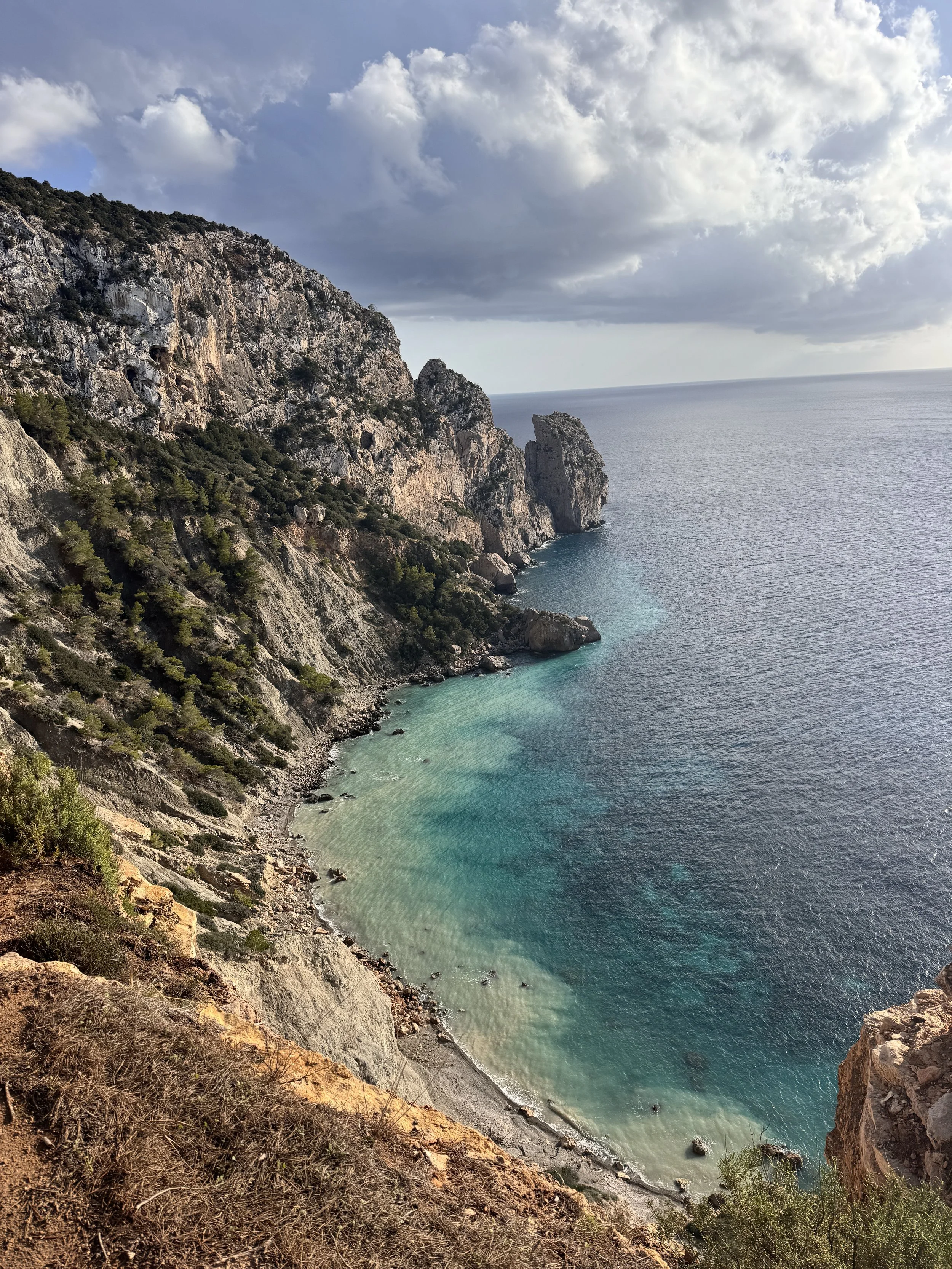 A rugged coastline with tall rocky cliffs covered in sparse greenery slopes down to a small, sandy beach. The water is a clear gradient of turquoise to deep blue, and the sky is partly cloudy with patches of sunlight.