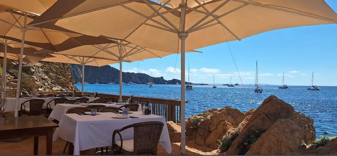 Ibiza restaurant. Outdoor restaurant seating area with white tables, chairs, and large beige umbrellas overlooking a rocky shoreline with sailboats on the water and a clear blue sky.