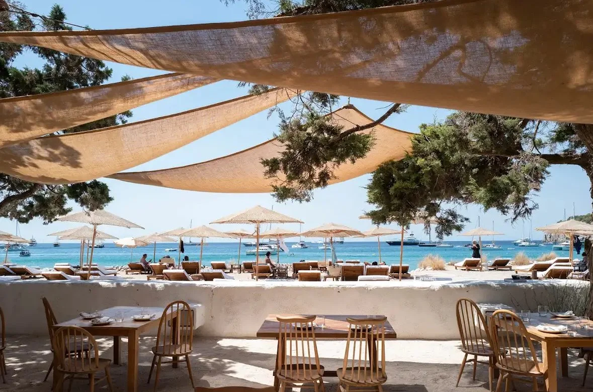 Jondal Ibiza. Beachside restaurant outdoor seating with wooden tables and chairs, shaded by large hexagonal sun sails, overlooking a sandy beach with lounge chairs and parasols, and boats anchored in the ocean.