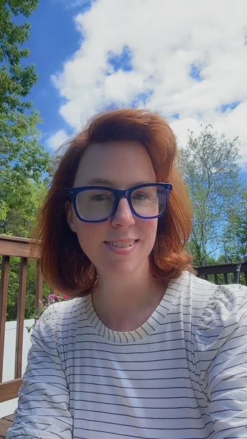 A woman with red hair and glasses taking a selfie outdoors on a sunny day with blue sky and clouds in the background.
