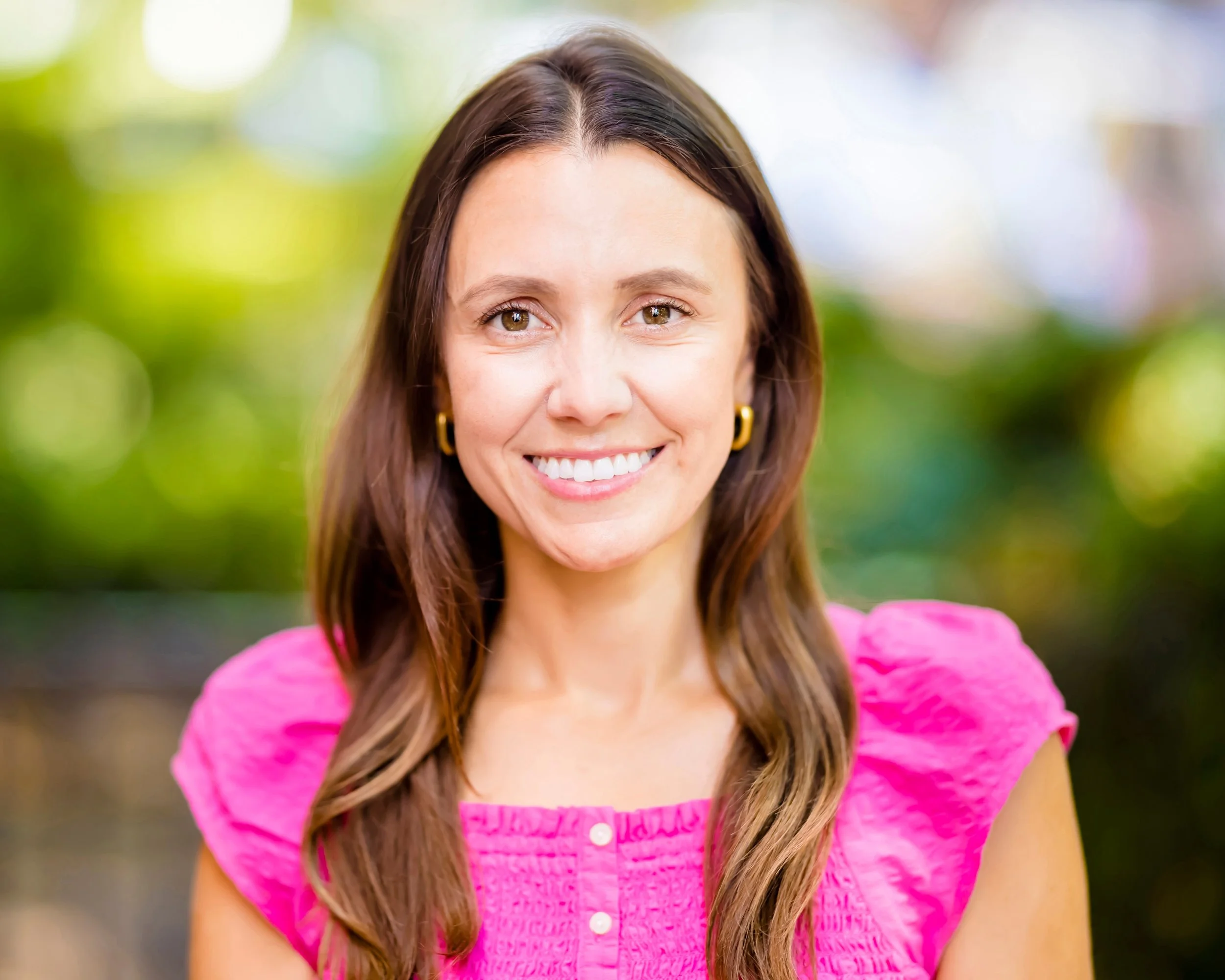 A woman smiling outdoors with a blurred green and colorful background, wearing a pink top and gold hoop earrings.