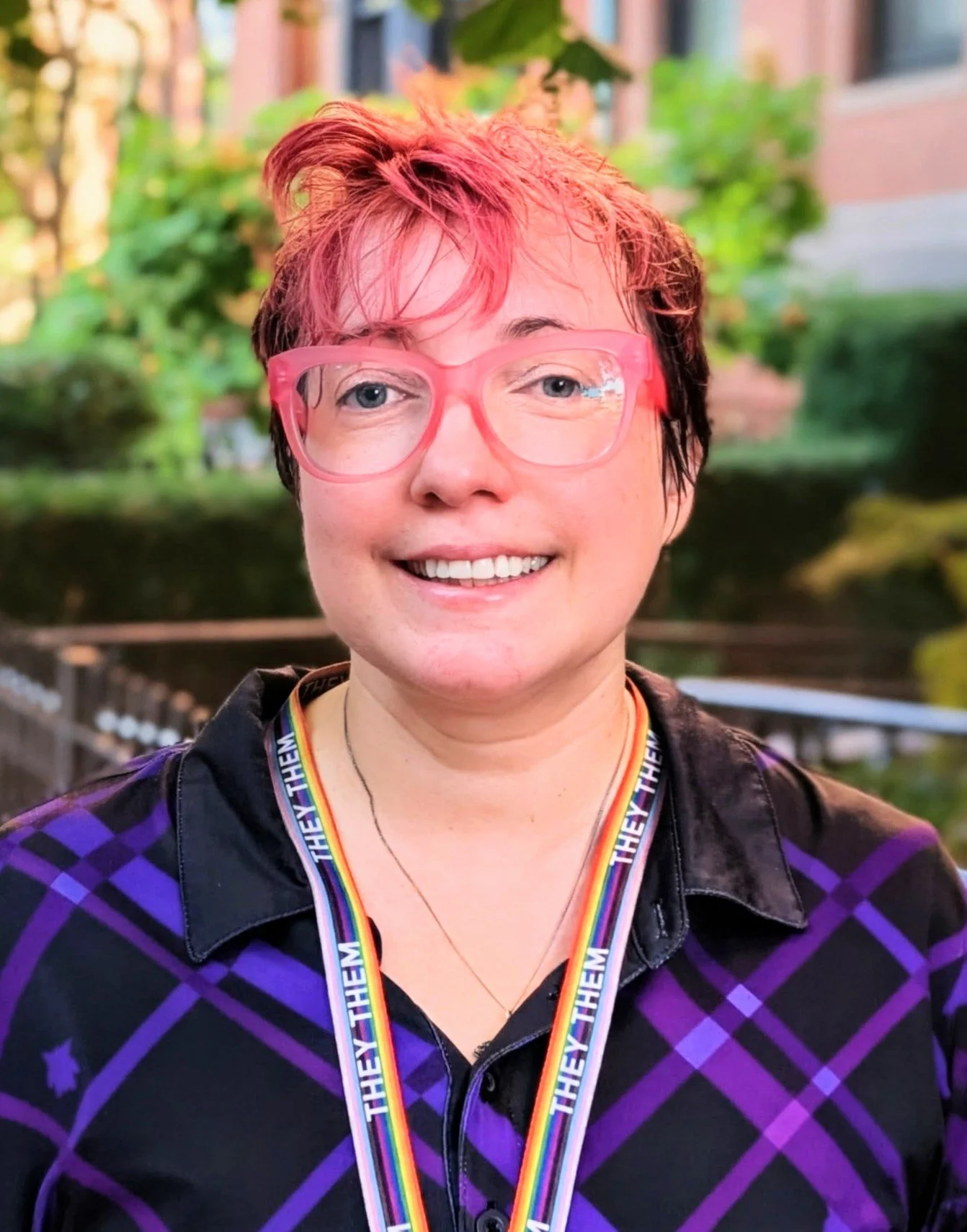 Person with short hair dyed pink with glasses, smiling outdoors, wearing a rainbow-colored medal ribbon that reads 'They Them' around their neck, wearing a black and purple checkered shirt with a collar, and standing in front of a blurred green background.