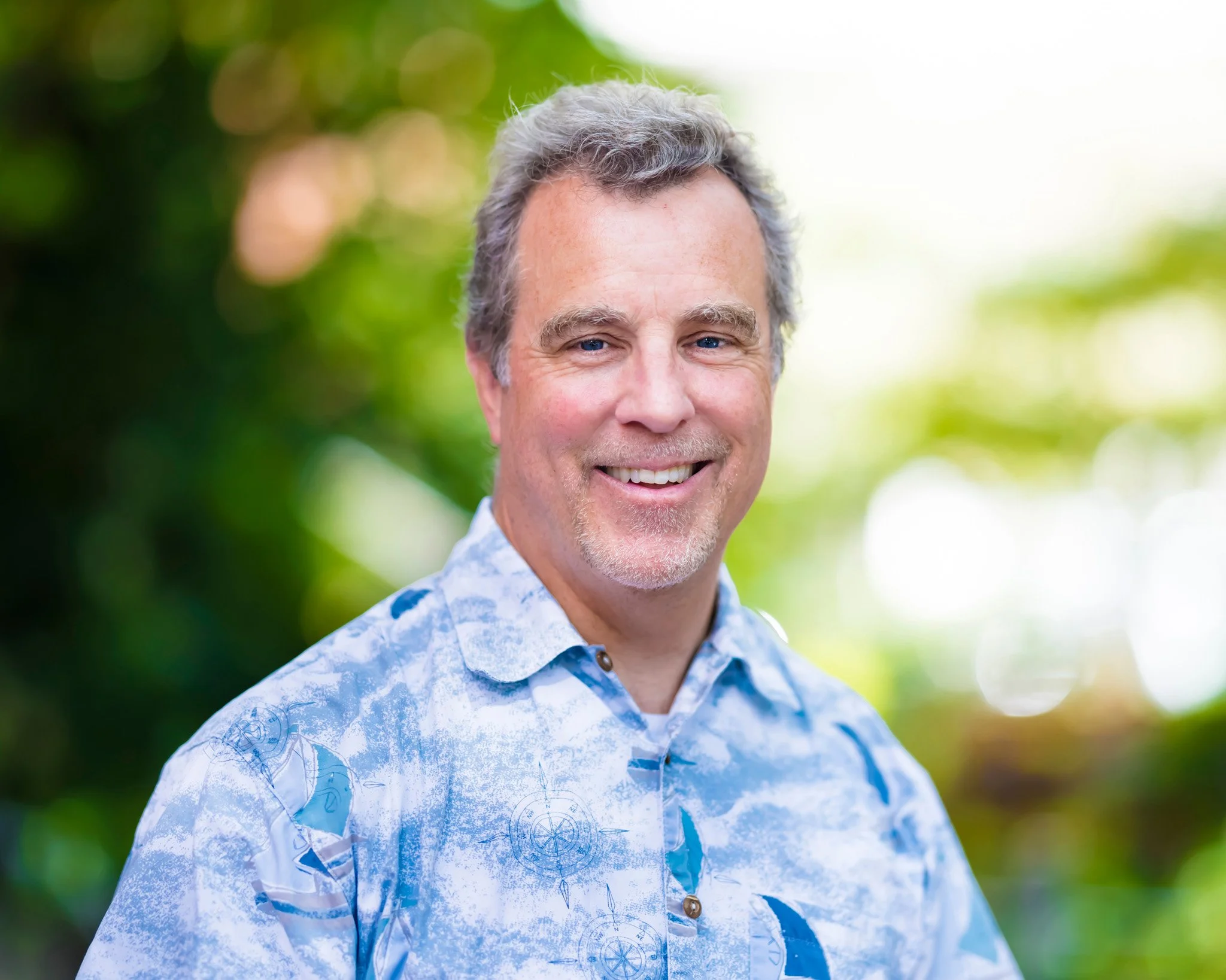 A smiling middle-aged man with gray hair and a goatee, wearing a blue patterned polo shirt, standing outdoors with a blurred green background.