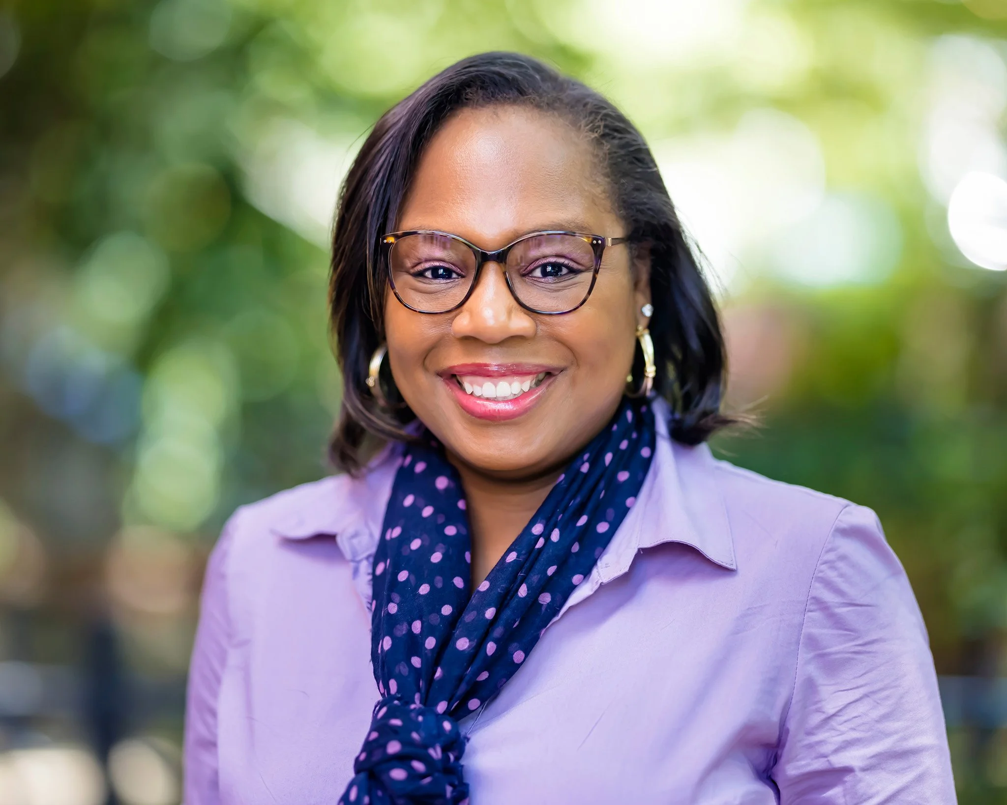 A woman with glasses, hoop earrings, and a blue polka dot scarf smiling outdoors with a blurred green background.