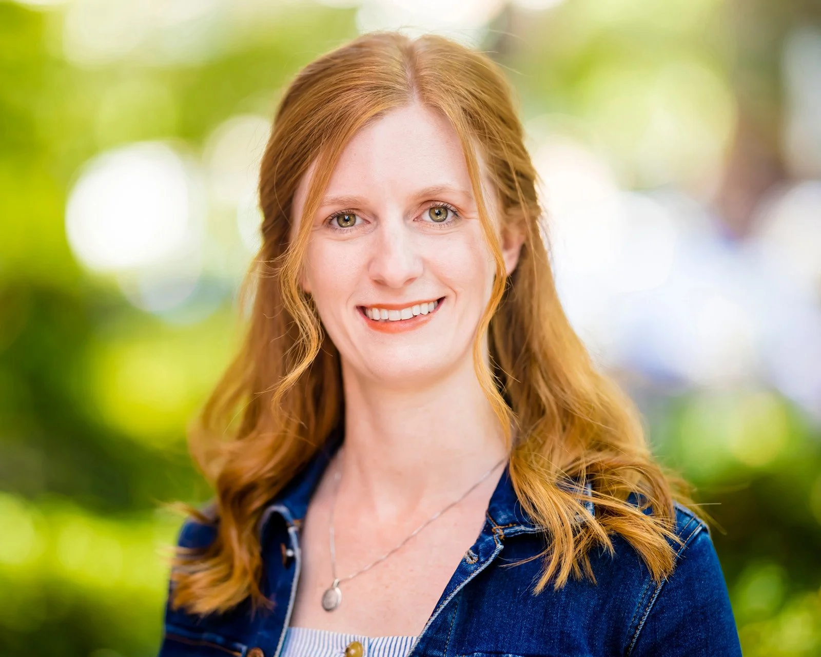Portrait of a woman with red hair, green eyes, wearing a denim jacket and a silver necklace, smiling outdoors with a blurred green background.