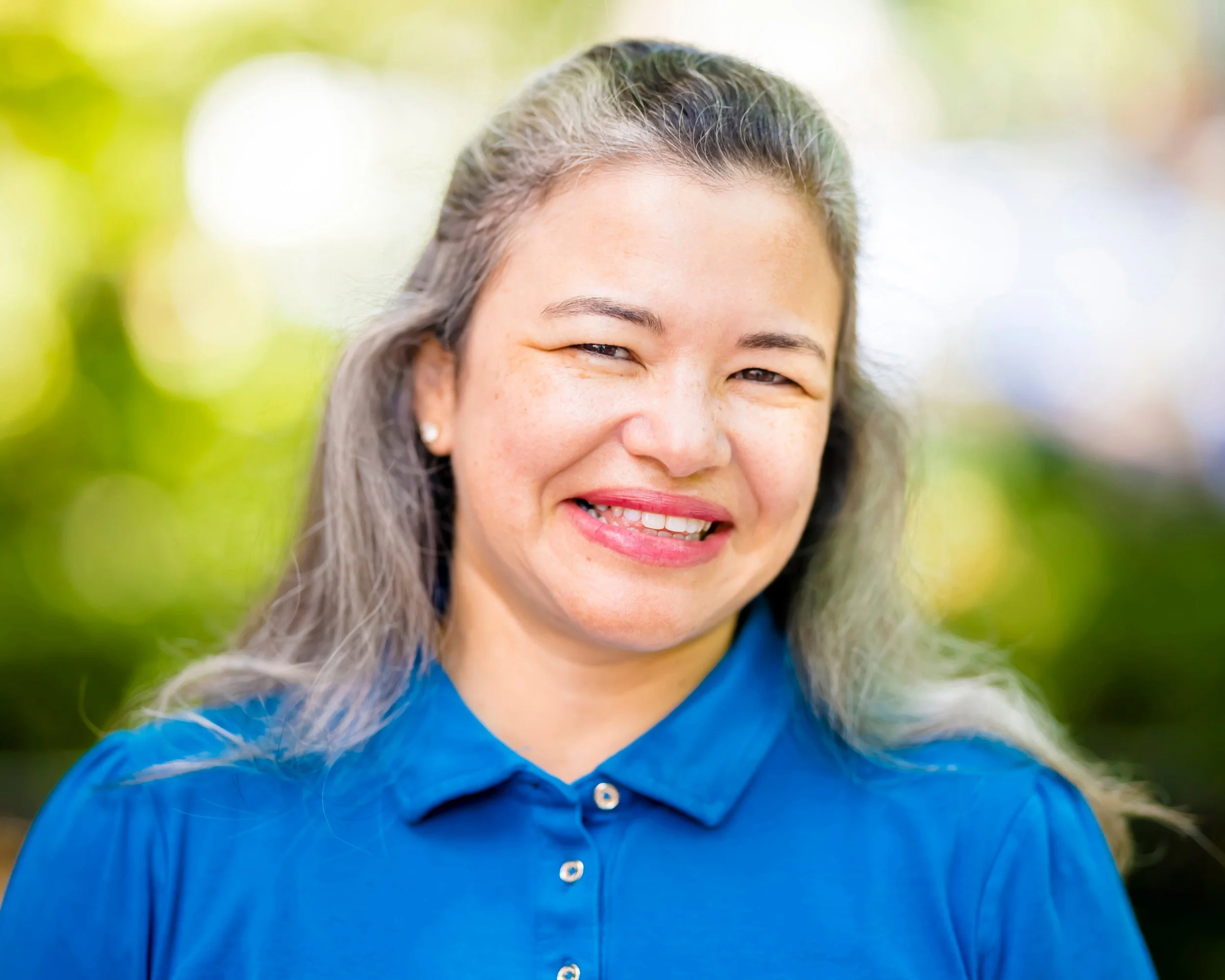 Close-up of a smiling woman with long gray hair wearing a blue collared shirt, outdoors with blurred greenery background.