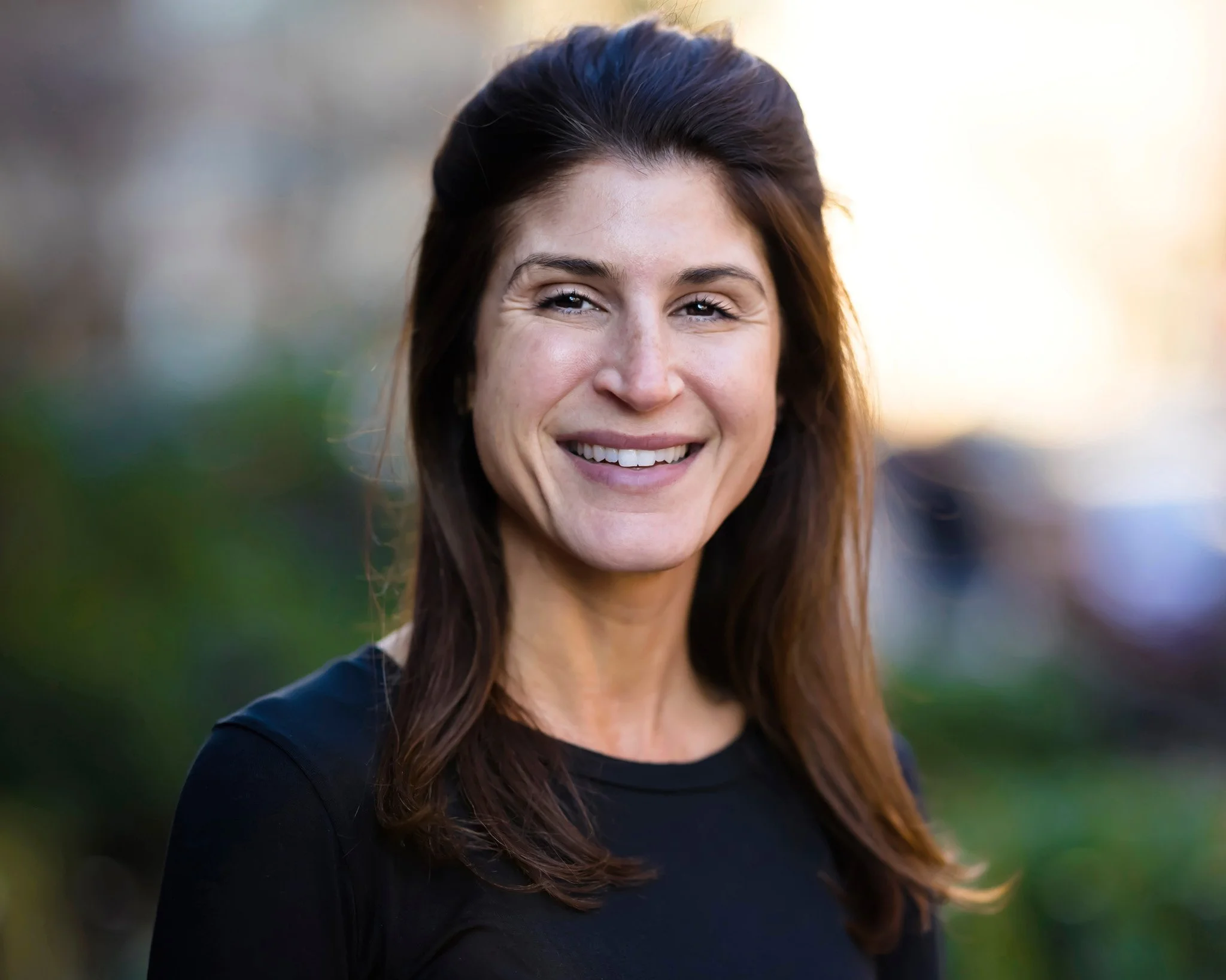 A woman with long brown hair styled back, smiling outdoors with blurred greenery in the background.
