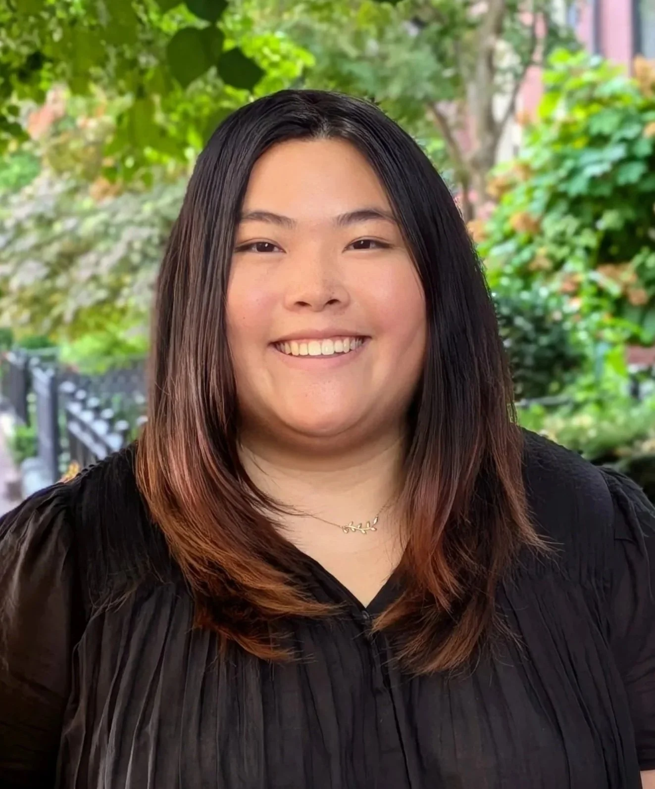 A young woman with long dark hair smiling outdoors with trees and greenery in the background.