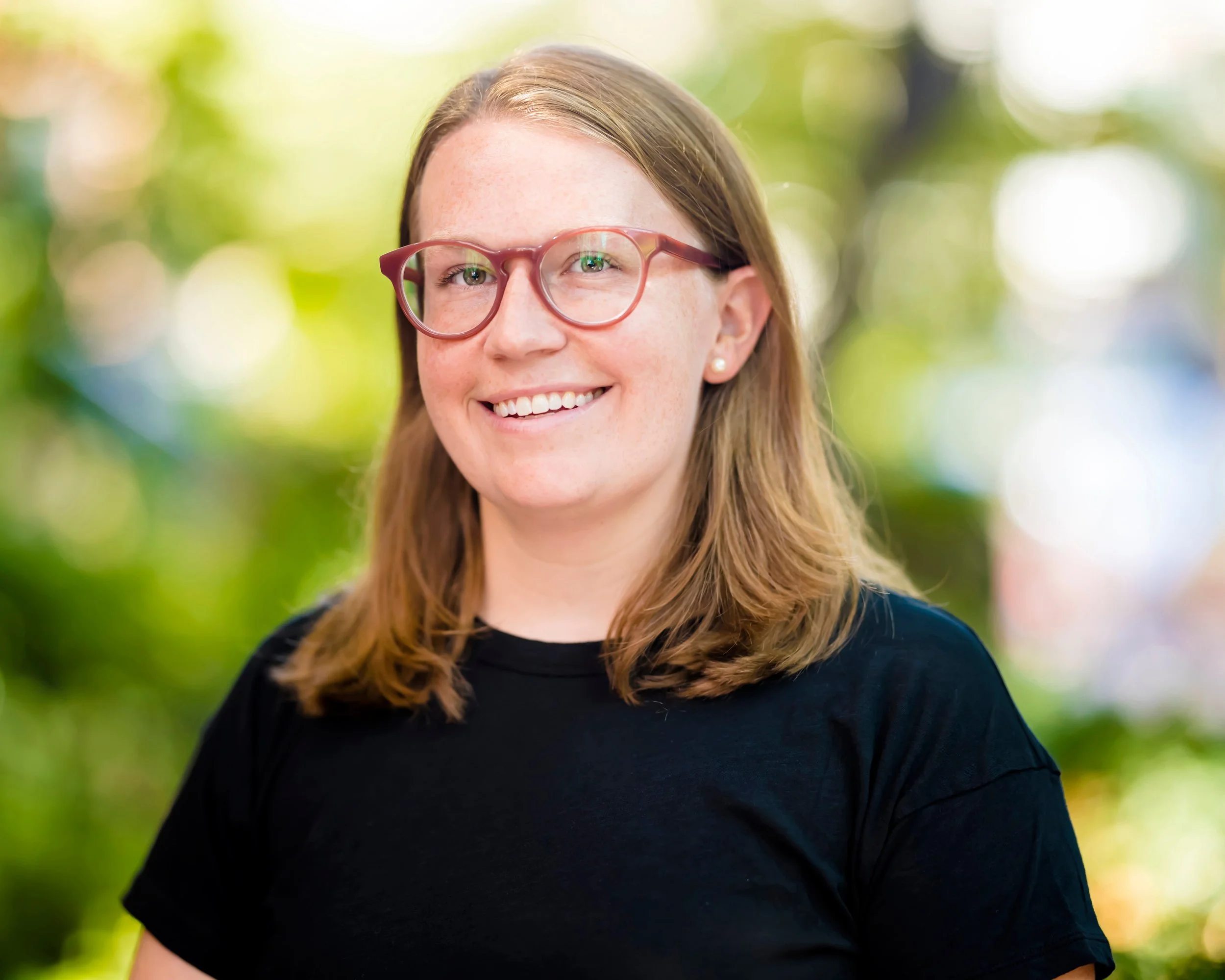 A young woman with glasses and shoulder-length brown hair smiling outdoors with a blurred green and yellow background.