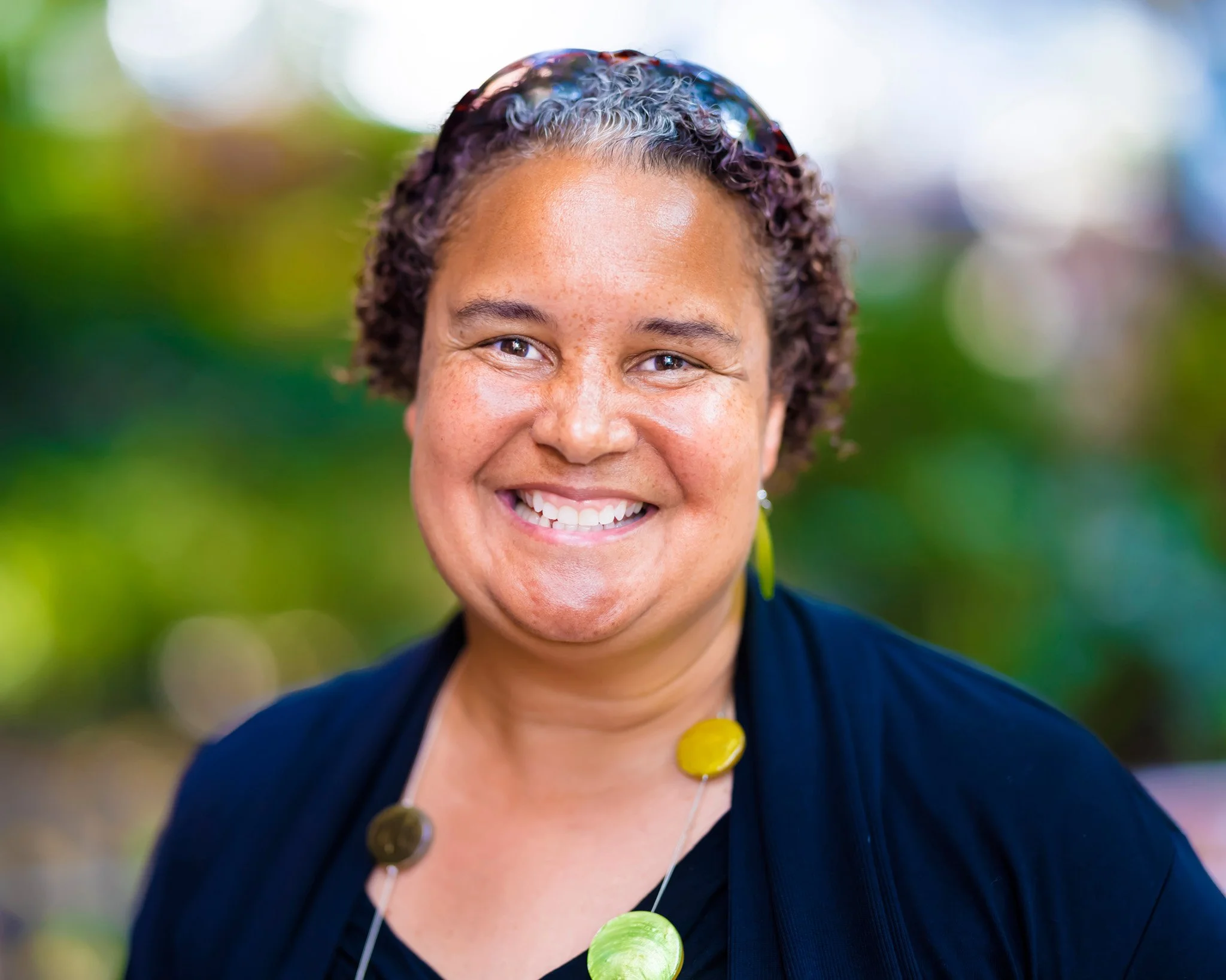 A woman with brown curly hair, smiling, wearing a black top, yellow earrings, and a colorful necklace, outdoors with a blurred green background.