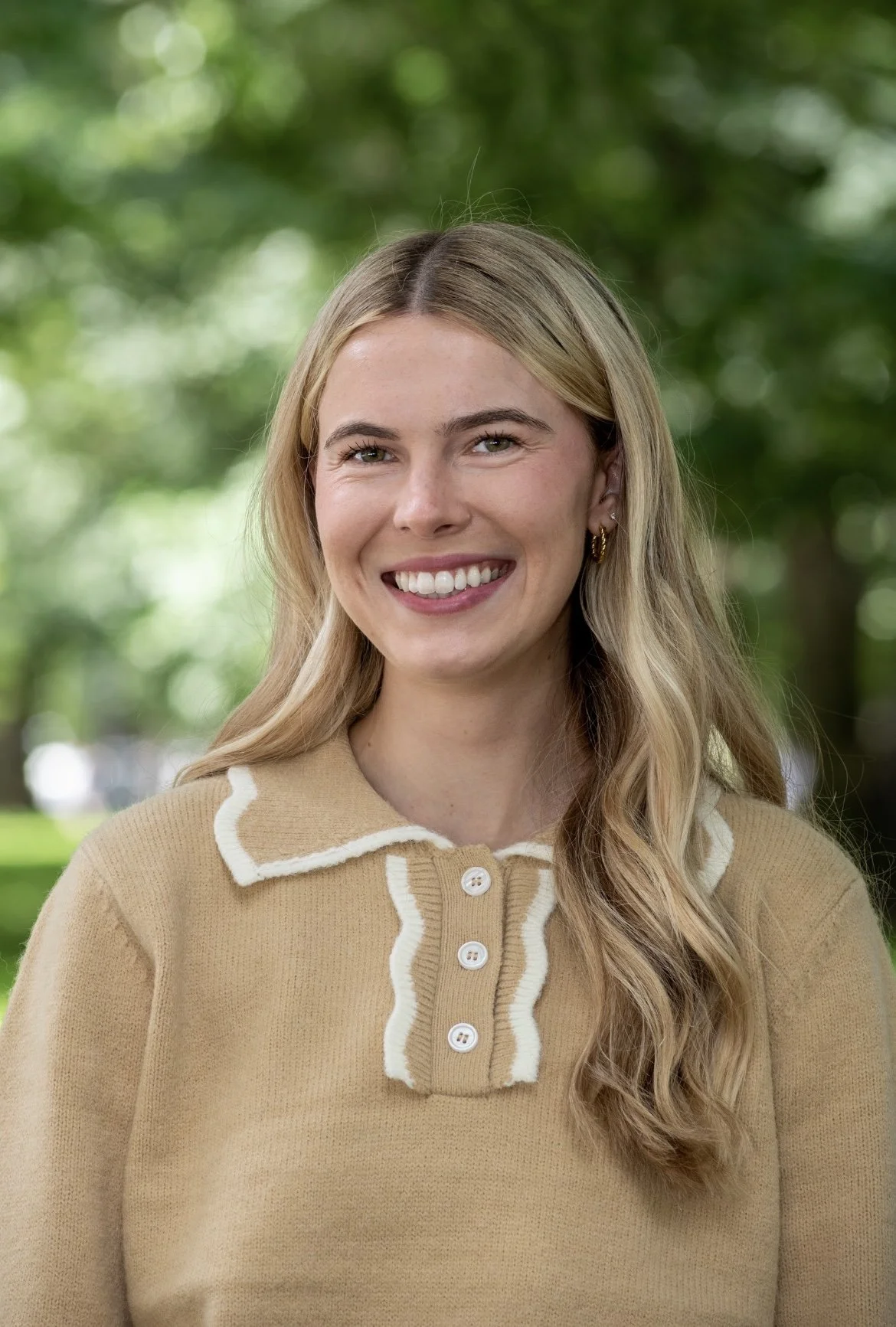 A young woman with long blonde hair, smiling, outdoors with green blurred trees in the background, wearing a beige sweater with white trim and buttons.