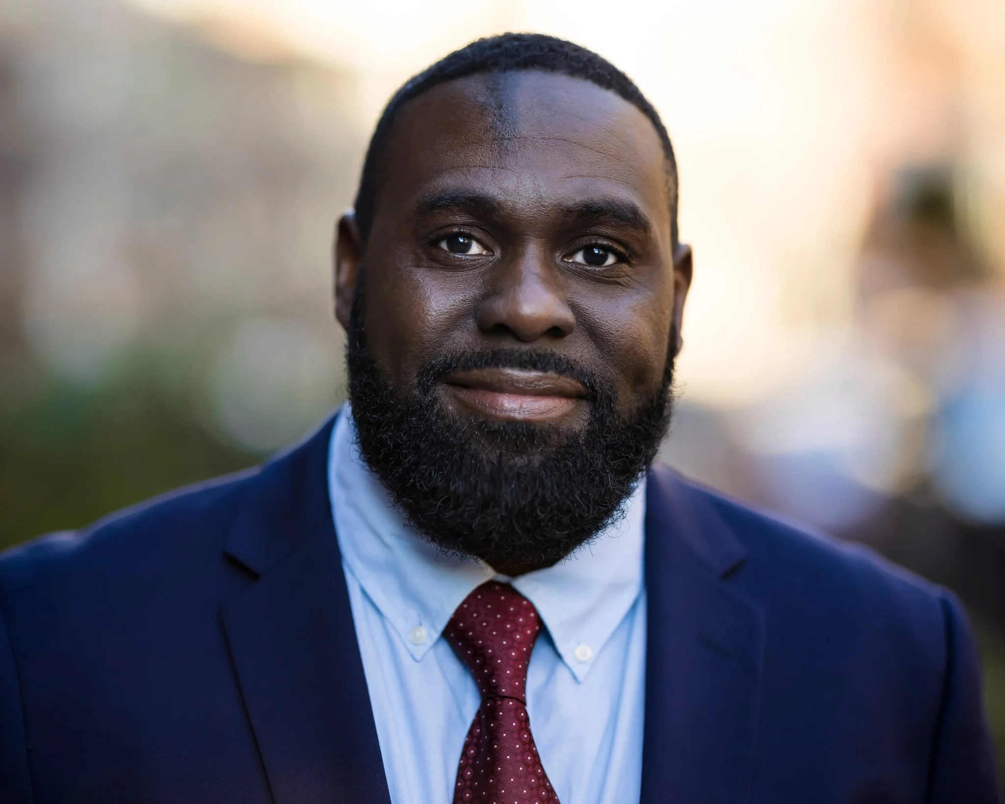 A man with a beard and short hair, wearing a navy blue suit, white shirt, and red tie, smiling outdoors during daylight.