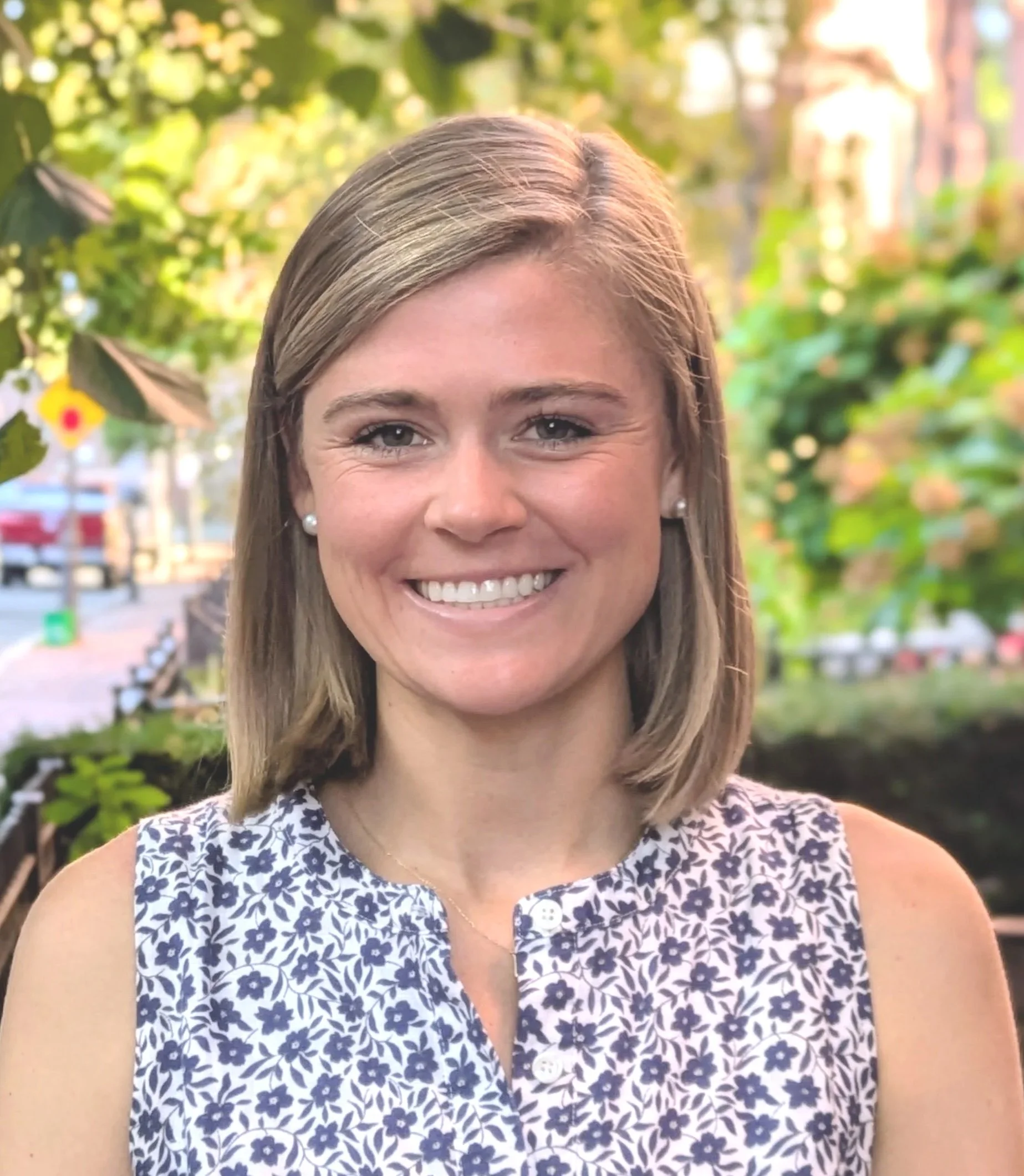 Smiling woman with straight blonde hair, pearl earrings, and a sleeveless blouse with a blue and white floral pattern, standing outdoors with green trees and blurred background.