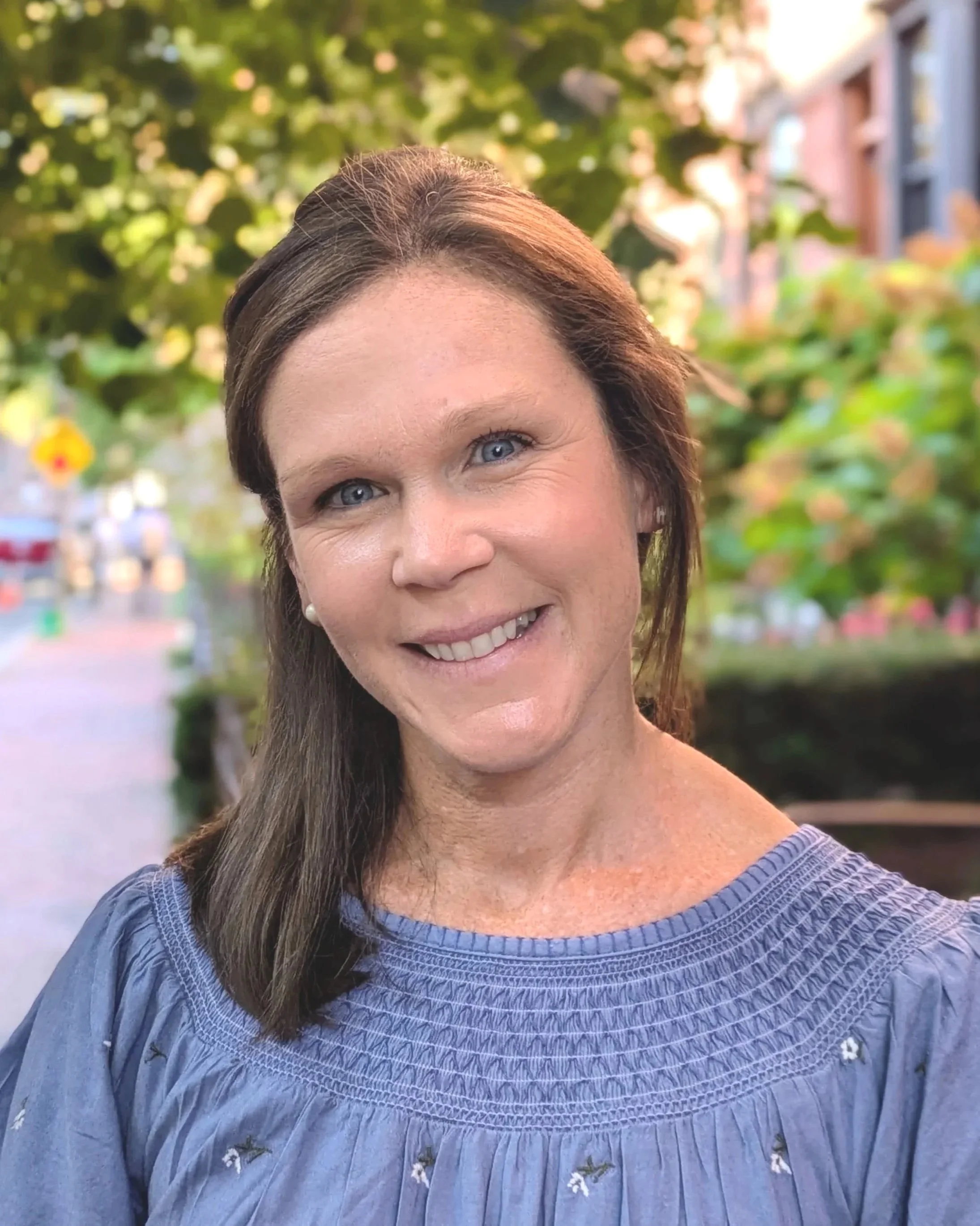 A woman with blue eyes and brown hair in a blue blouse smiling outdoors with trees and buildings in the background.