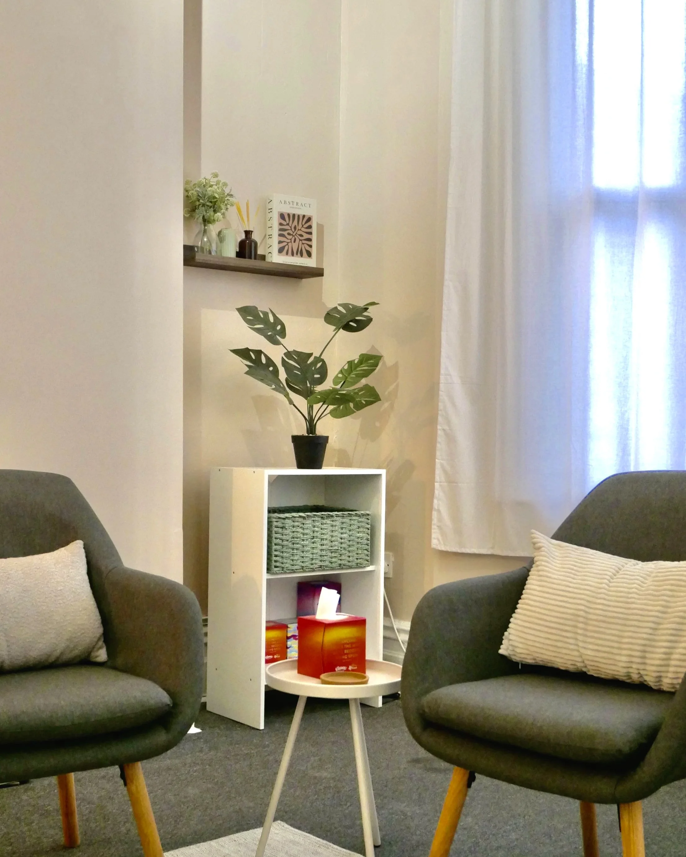 Living room with two gray armchairs, beige cushions, a white side table with tea boxes, a white shelf with a potted plant and decorative items, and a wall shelf with books and small decor, near a window with white curtains.