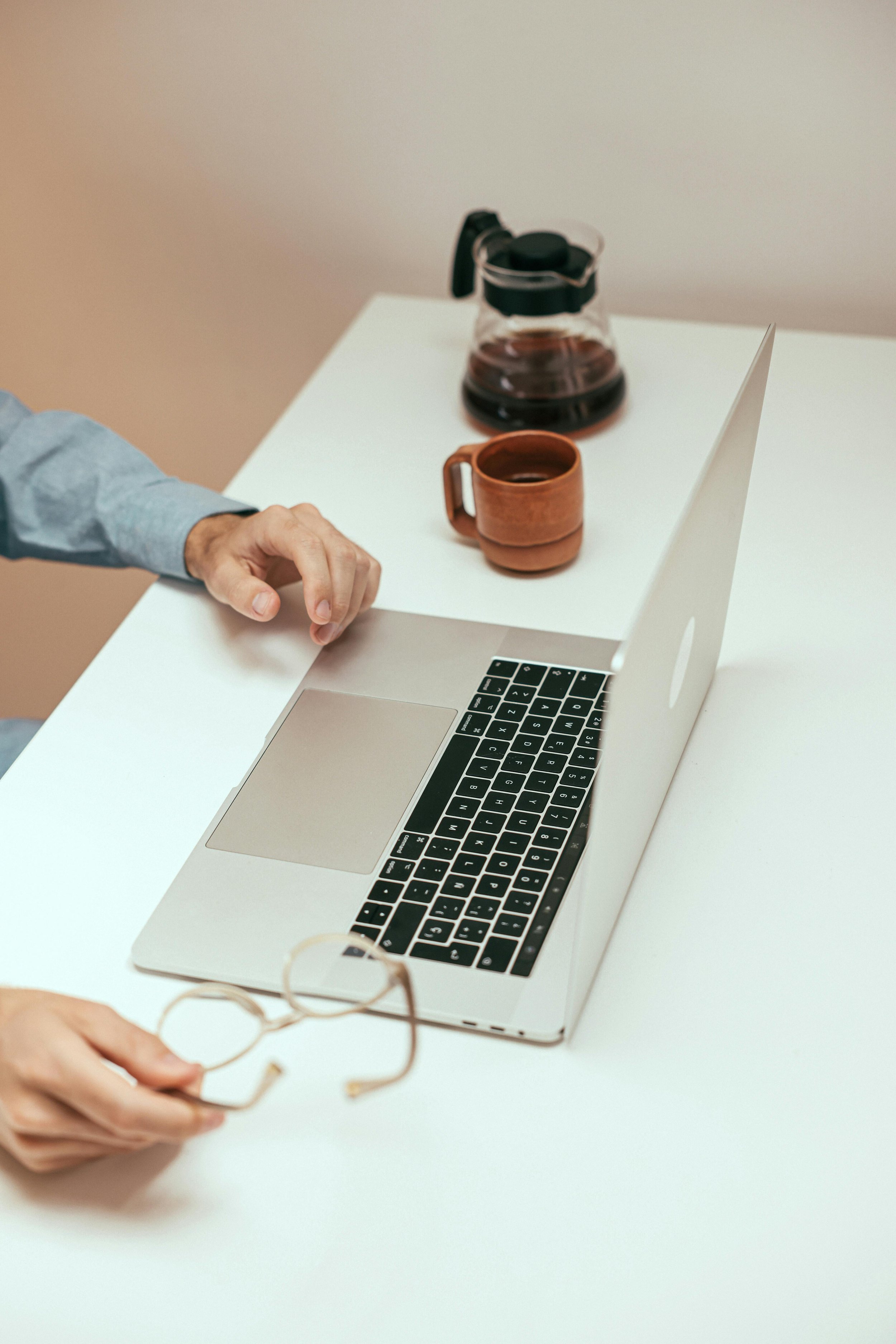 A hand holding glasses with a thin wooden frame, a laptop on a white table, a glass coffee pot, and a brown ceramic mug.
