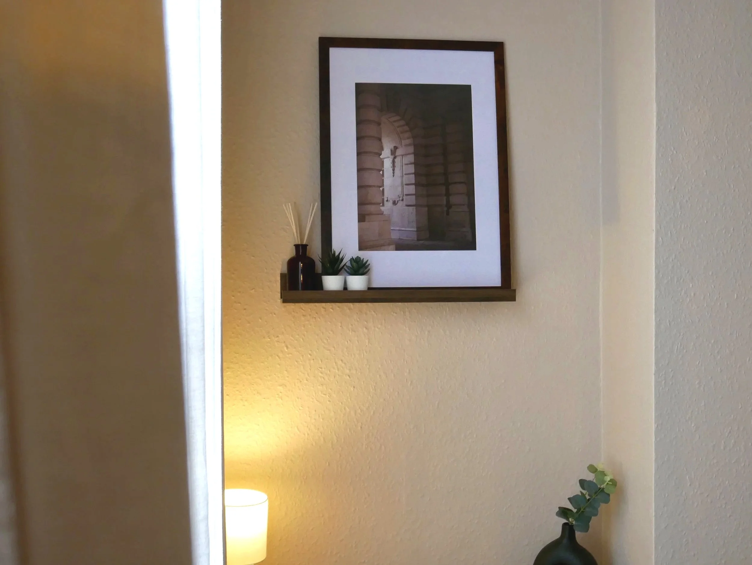 Decorative wall arrangement with a framed black-and-white architectural photograph, small potted plants, and vases on a small shelf, with a lamp and green plant in the corner.