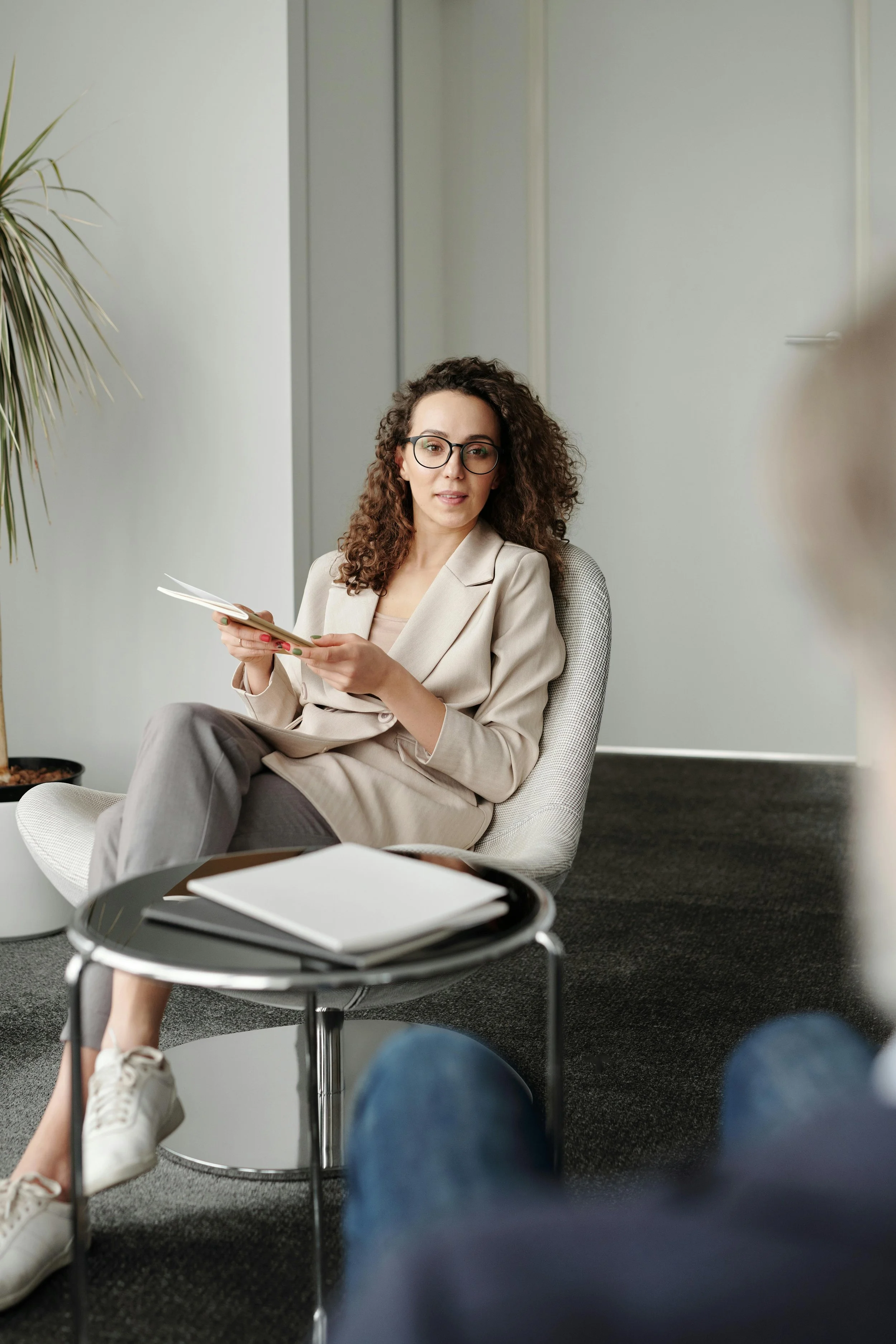 A woman with curly brown hair and glasses sitting in a modern office. She wears a beige blazer, gray pants, and white sneakers, and is holding a notebook. In the foreground, there is a black table with a closed white notebook on it. The background features a white wall and a tall potted plant.