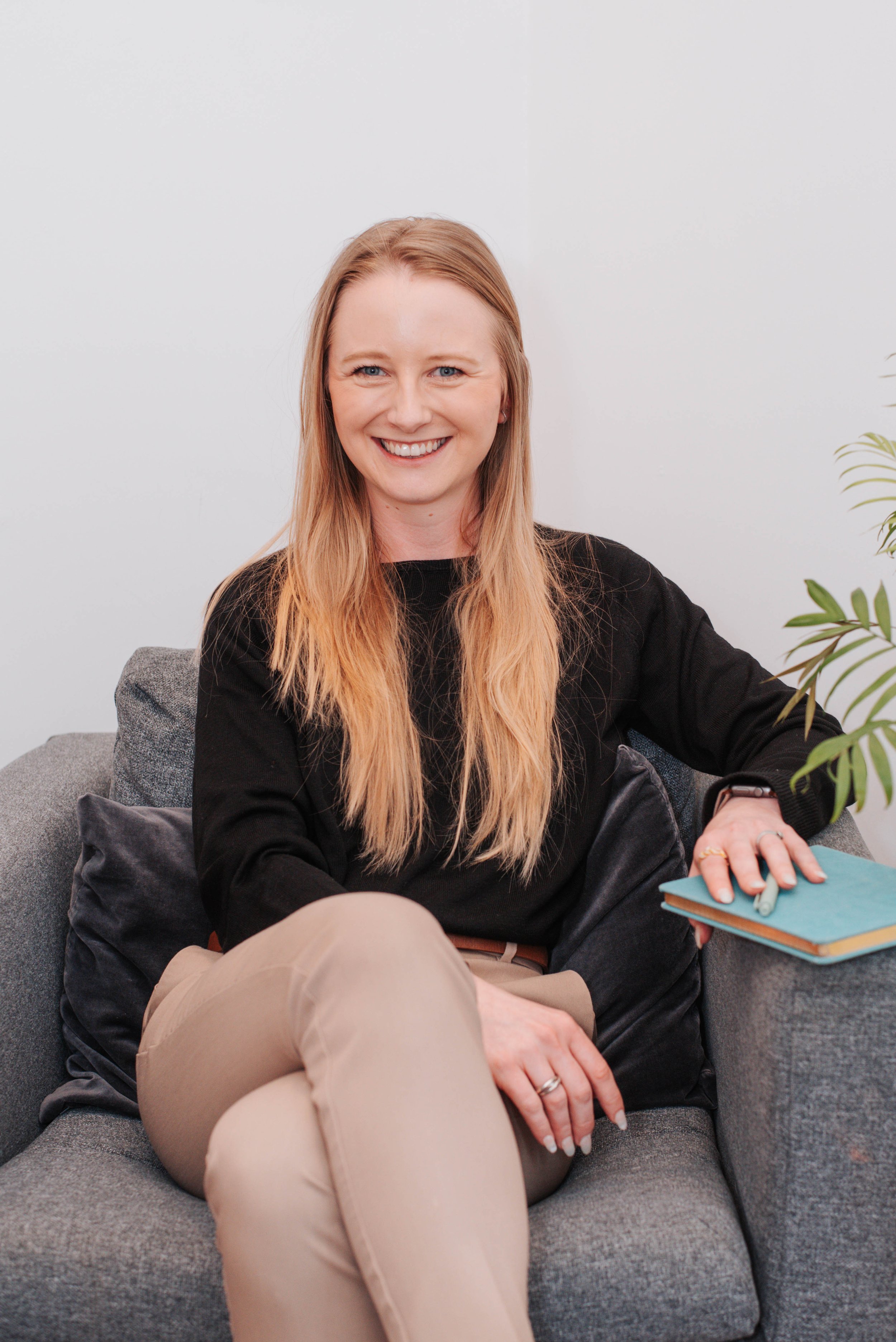 A young woman with long blonde hair and a black shirt sitting on a gray couch in a room with white walls, smiling at the camera. She has a notebook and pen on her lap and a plant visible on the right side of the image.