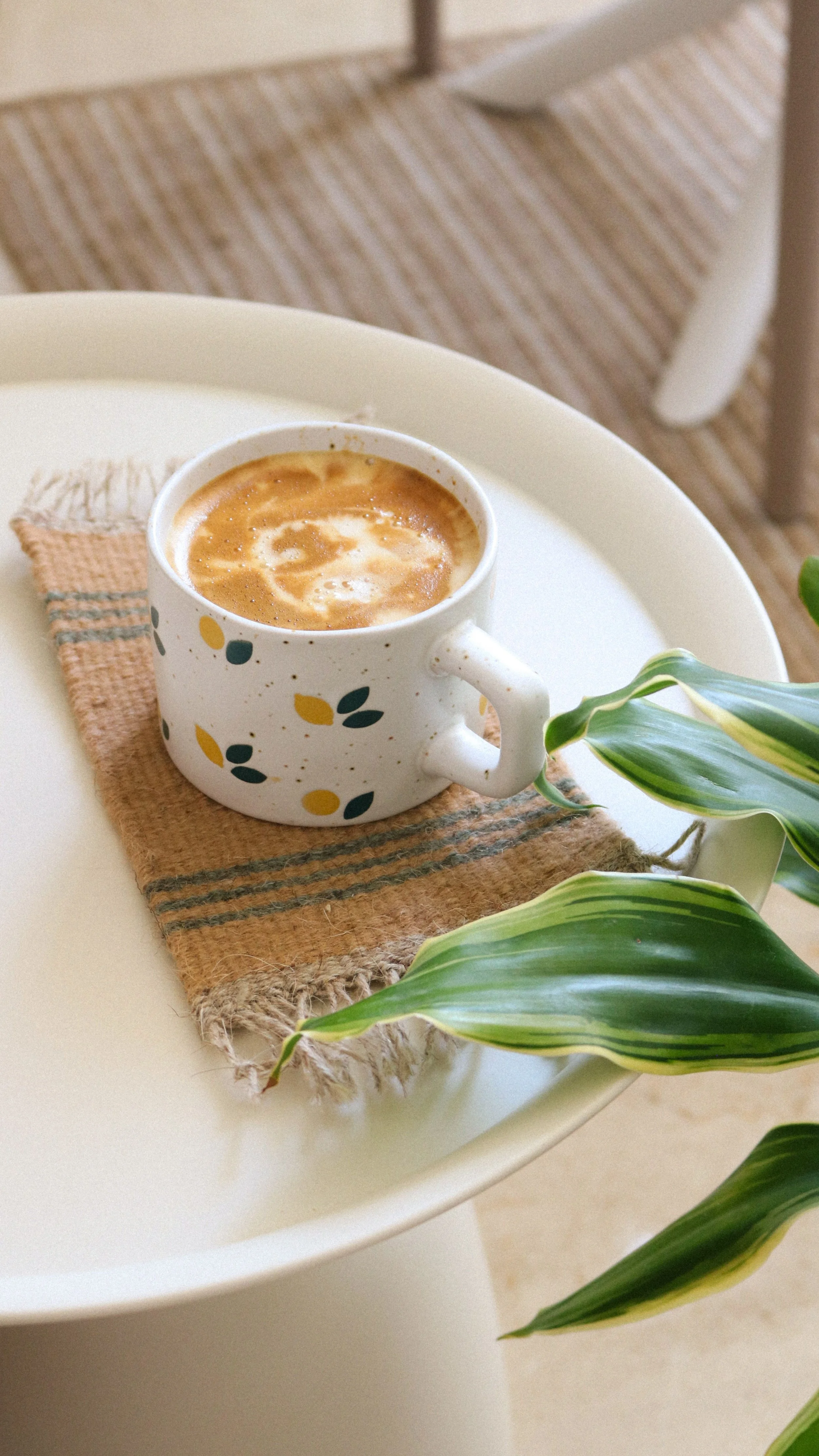 A decorative mug containing coffee with foam art, placed on a woven mat on a white tray. A green leafy plant is partially visible in the foreground.