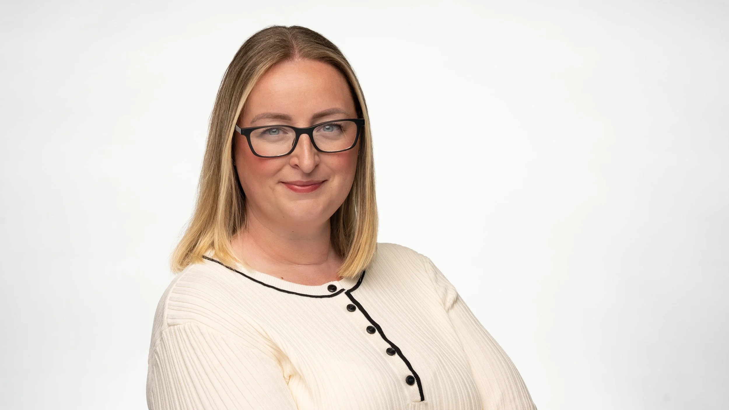 A woman with shoulder-length blonde hair wearing large black glasses and a cream-colored sweater with black trim, smiling softly against a plain white background.