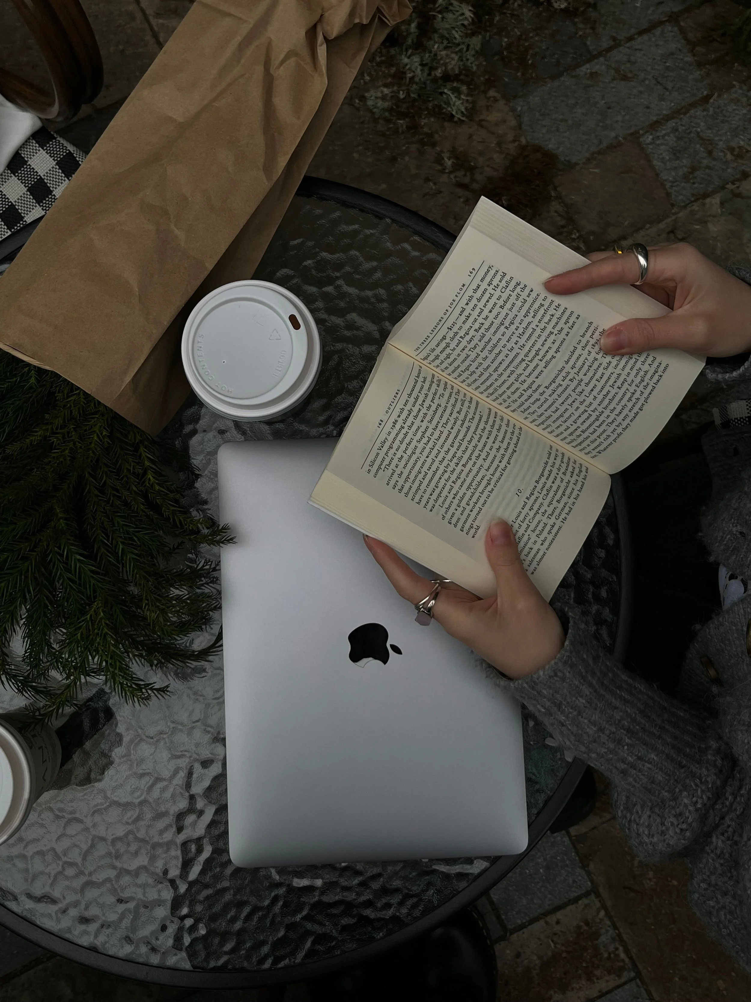 A person reading a book outdoors at a table with a laptop, disposable coffee cup, and paper bag.