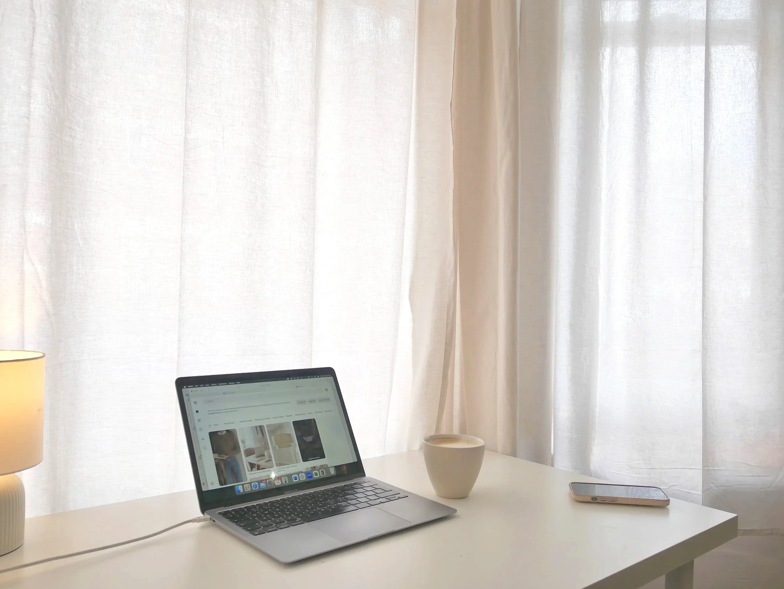 A white desk with a laptop, a smartphone, a white mug, and a small lamp in front of beige curtains.