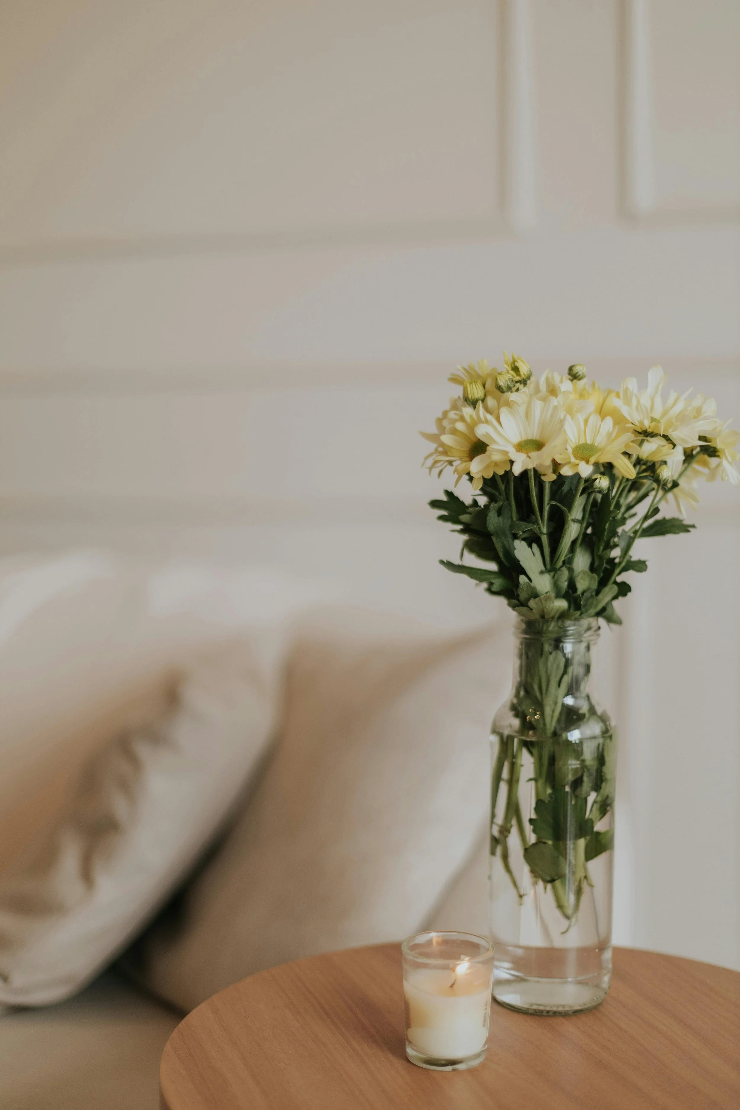 A vase of white and yellow daisies and a lit candle on a wooden table in a cozy room.