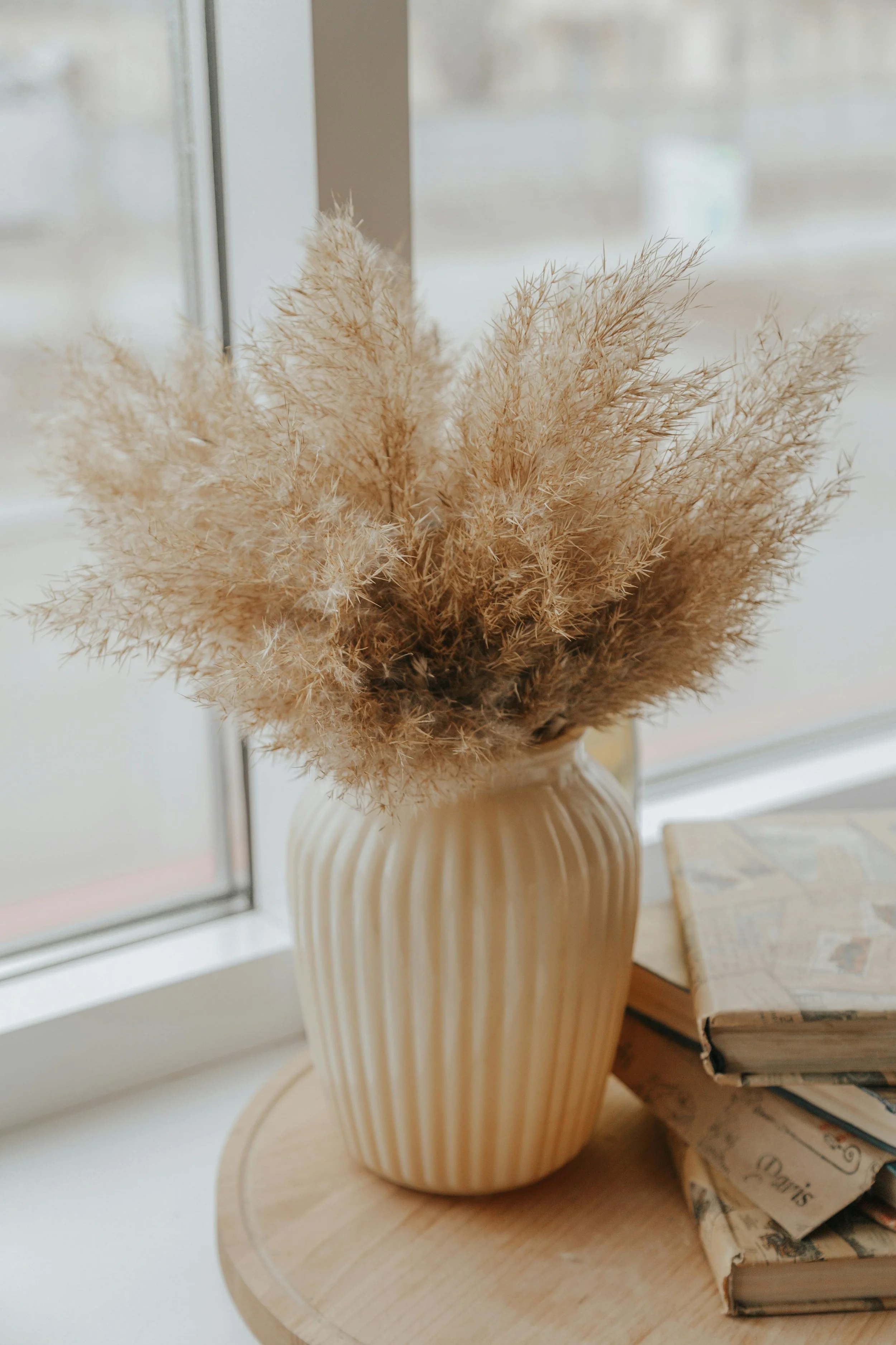 A beige ceramic vase with vertical ridges holding dried pampas grass, placed on a round wooden table near a window, with a stack of old books beside it.