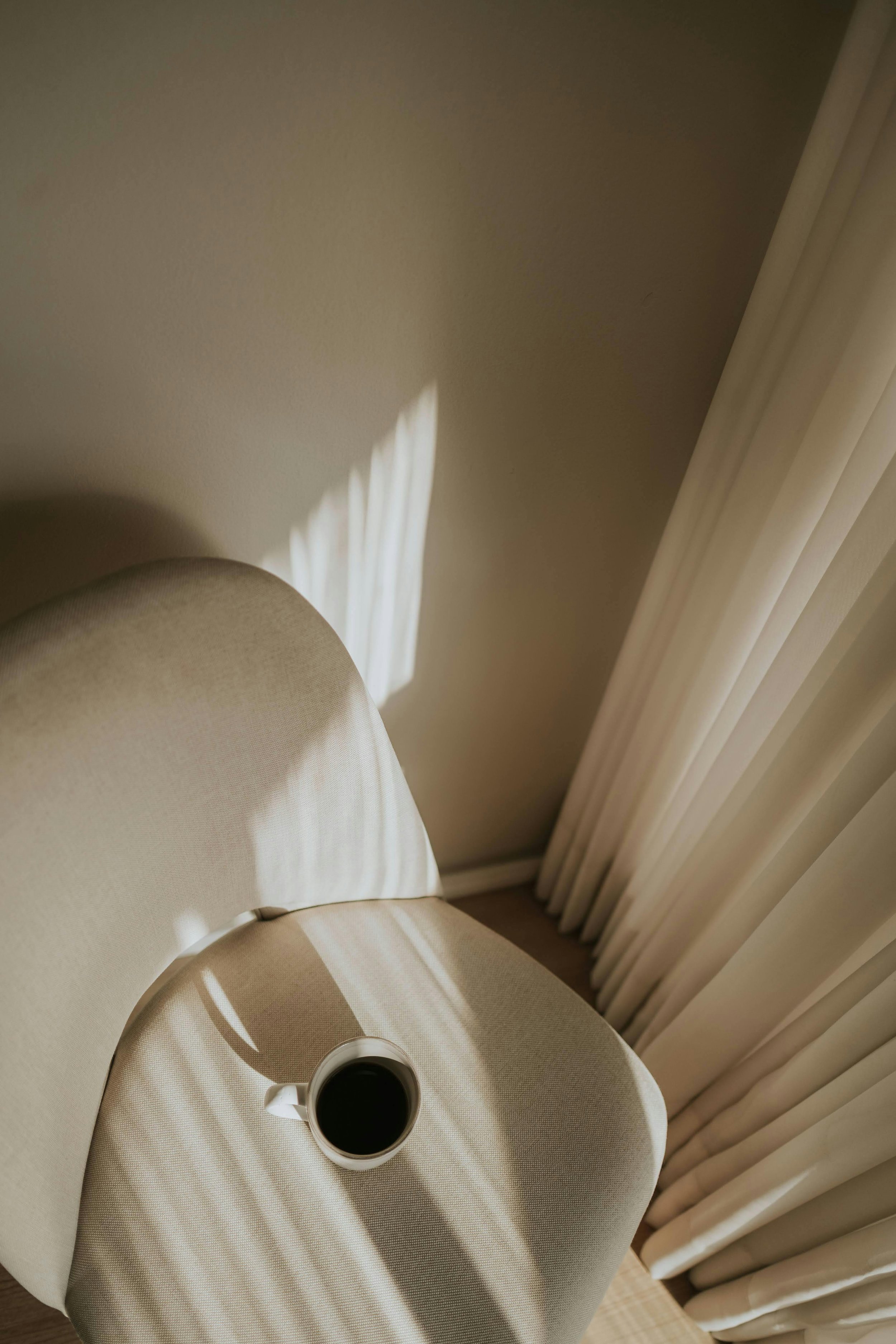A beige chair with a round white side table on which a white mug filled with black coffee sits, with sunlight and shadow stripes cast on the wall and curtain in the background.