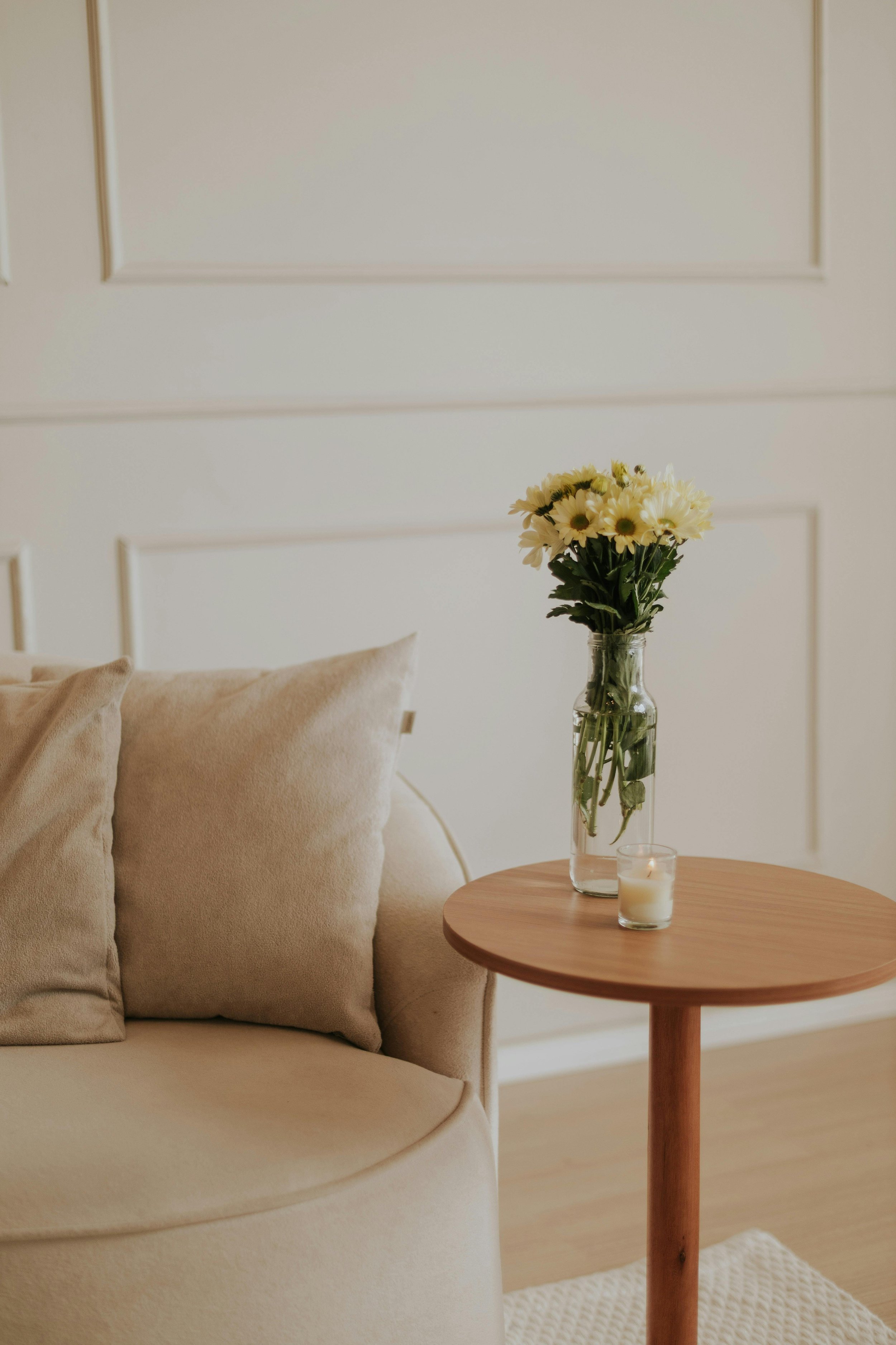 A cozy living room corner with a beige sofa, beige pillows, a small round wooden side table holding a glass vase of white and yellow flowers and a lit candle, and a textured white rug.