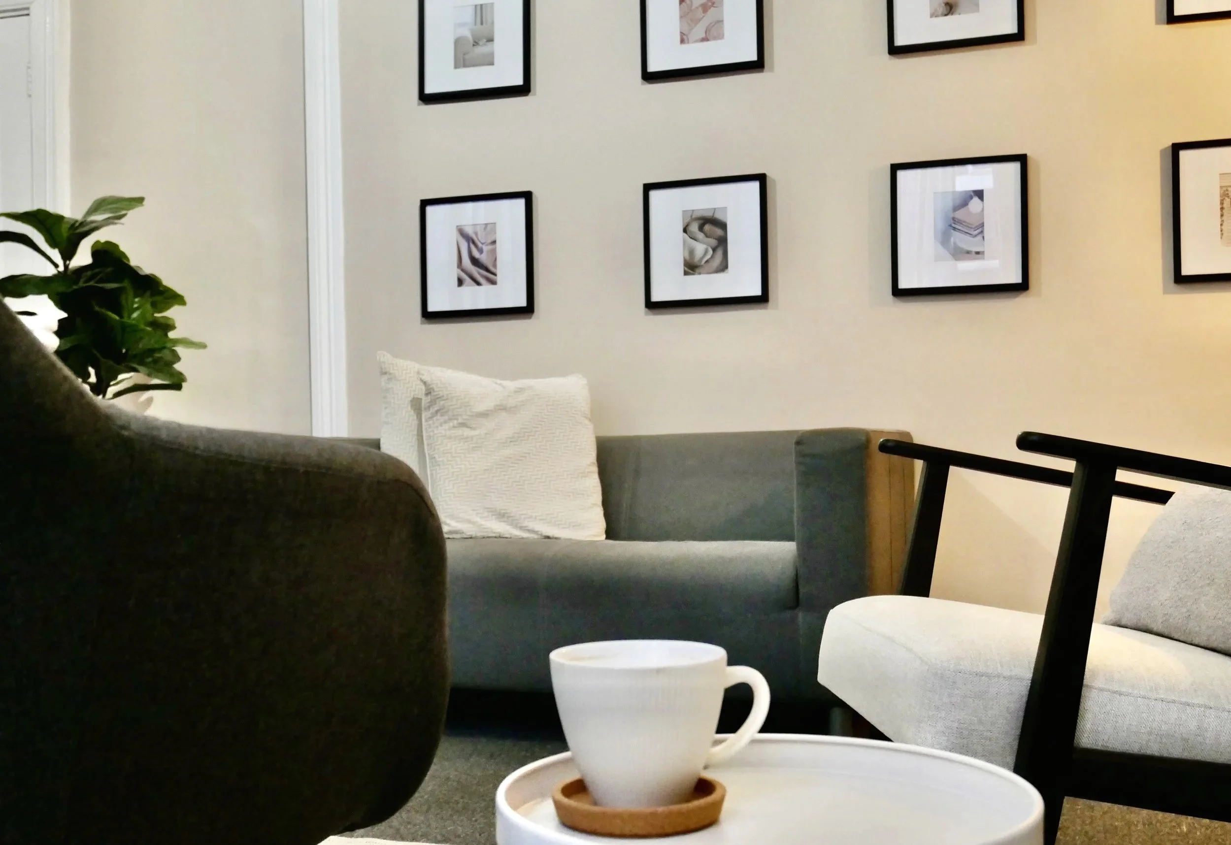 Living room with gray sofa, white pillow, framed artwork on beige wall, green plant, white coffee cup on tray, gray chair.
