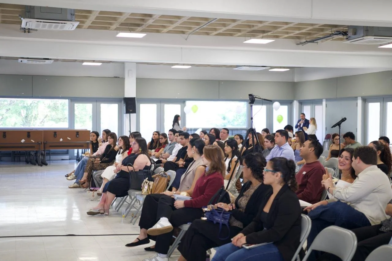 People sitting in a conference room listening to a presentation, with some using notebooks and others talking. Windows with green scenery outside, and balloons inside the room.