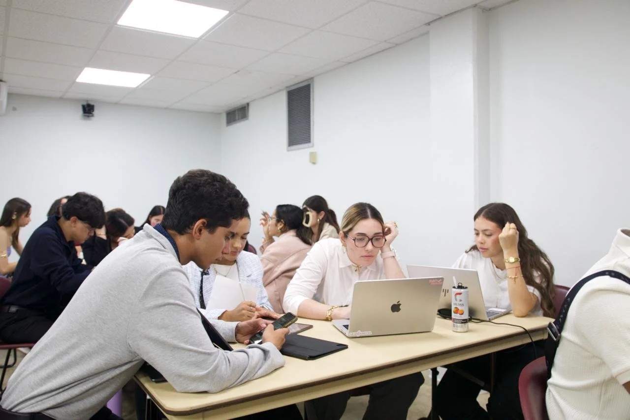 Young adults sitting at a table in a classroom or meeting room, using laptops and smartphones, with others in the background working or listening.