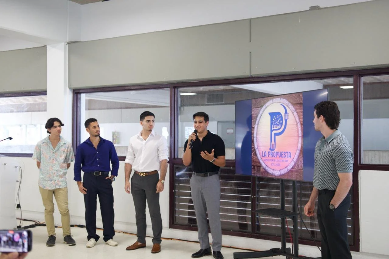 Five young men standing in a row, with one speaking into a microphone. A large digital screen displays a logo with the text 'La Propuesta' and an illustration of a fork and knife. The setting appears to be an indoor presentation or event space.