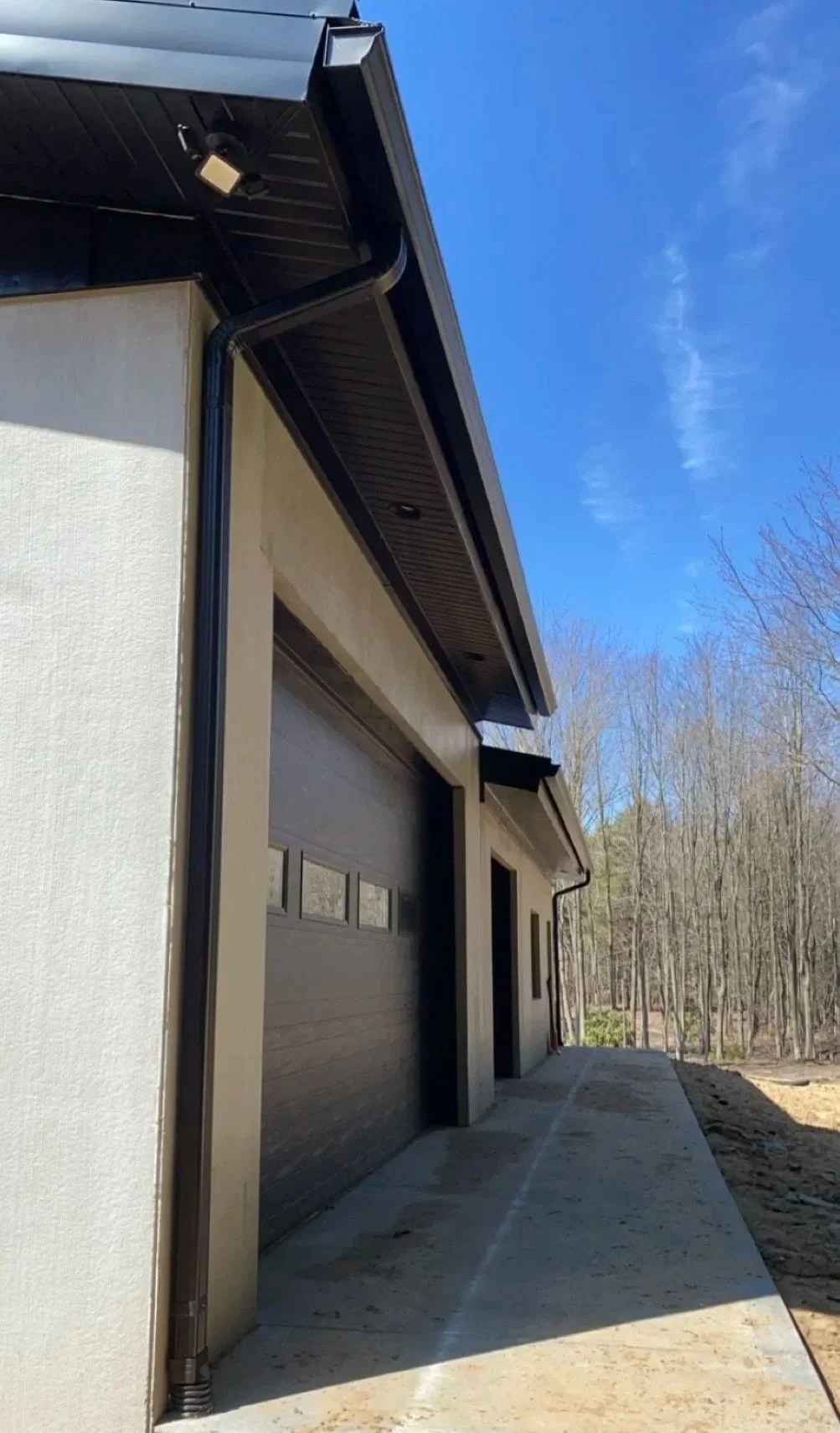 Front of a modern house with a garage door, surrounded by a concrete driveway and trees in the background. The house has black gutters and fascia, with a clear blue sky above.