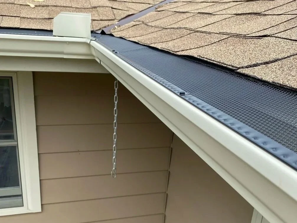 Close-up of a house's gutter system with a chain hanging from it, beige exterior siding, and a window, with a shingle roof in the background.