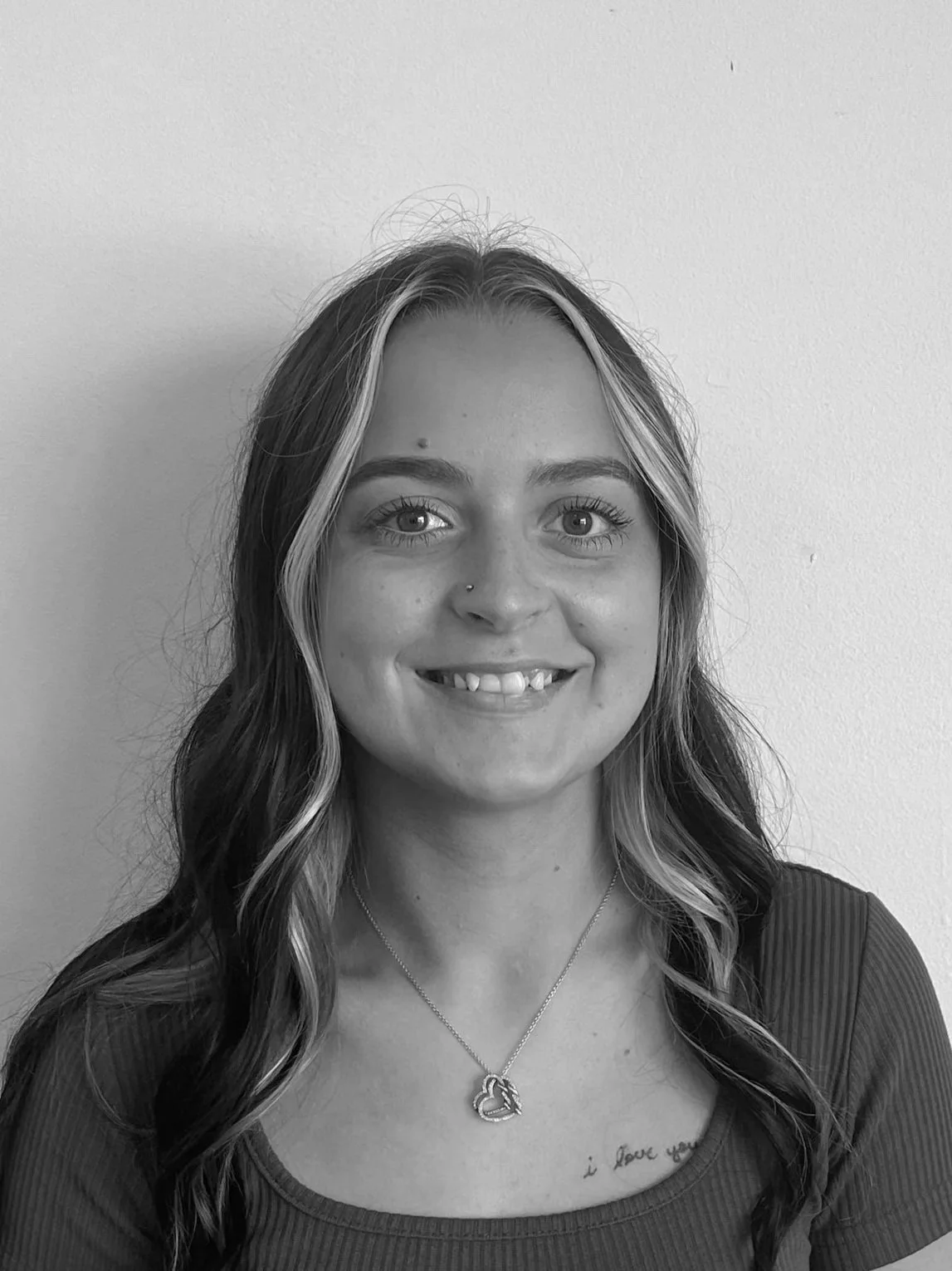 Black and white portrait of a smiling woman with shoulder-length wavy hair, wearing a dark top and a lacy black cardigan, standing against a plain light-colored wall.