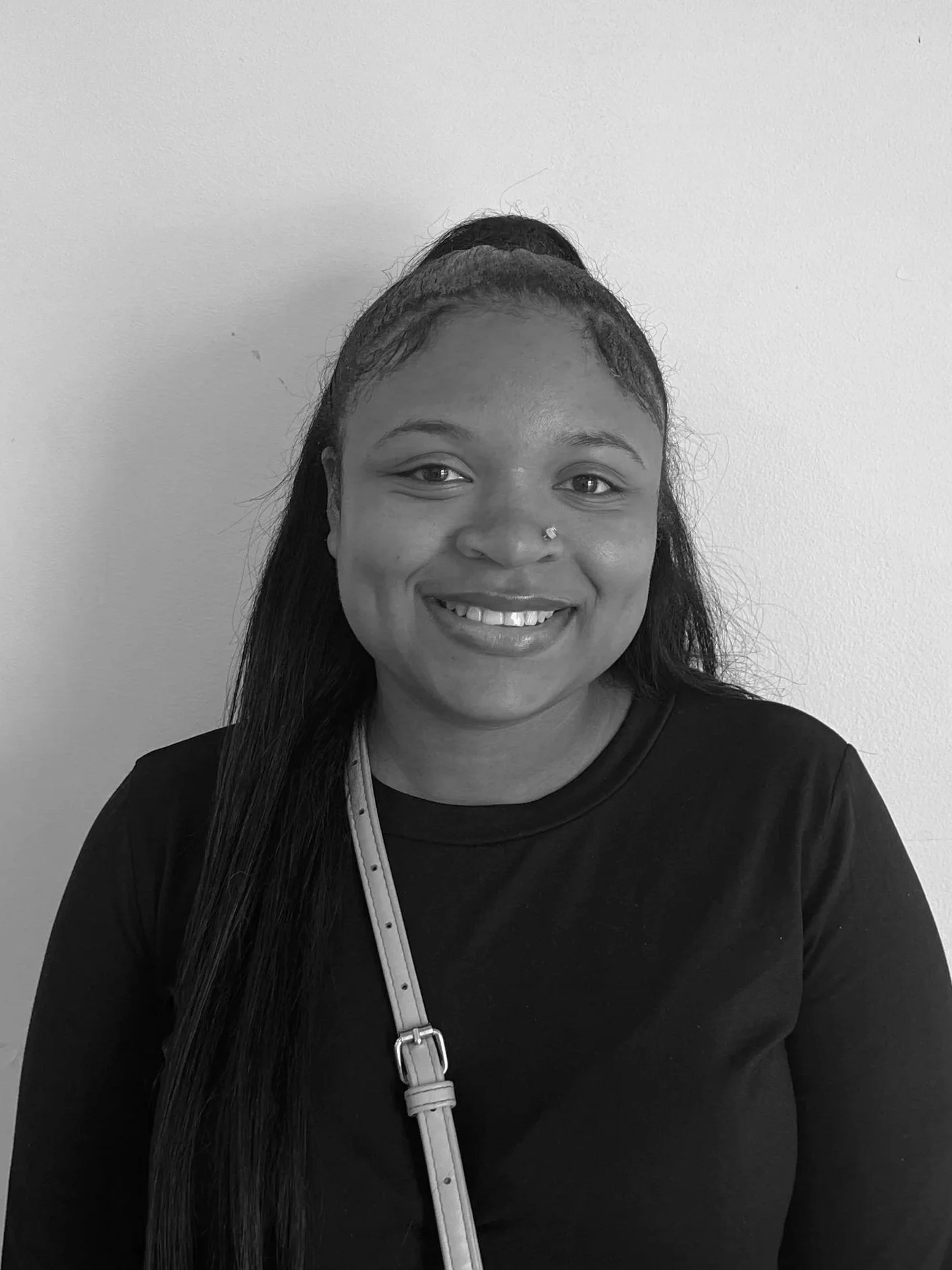 A smiling young woman with shoulder-length blonde hair, wearing a patterned sleeveless jacket over a dark t-shirt, standing against a plain white wall.