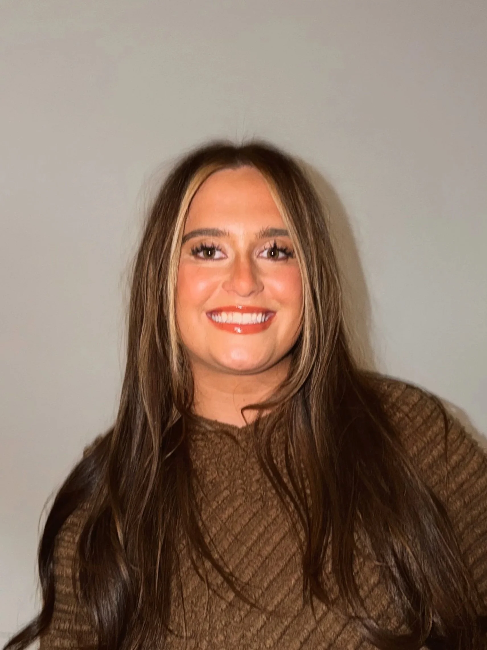 A woman with long wavy brown hair holding a Canon camera, standing outdoors near columns with a grassy area in the background.