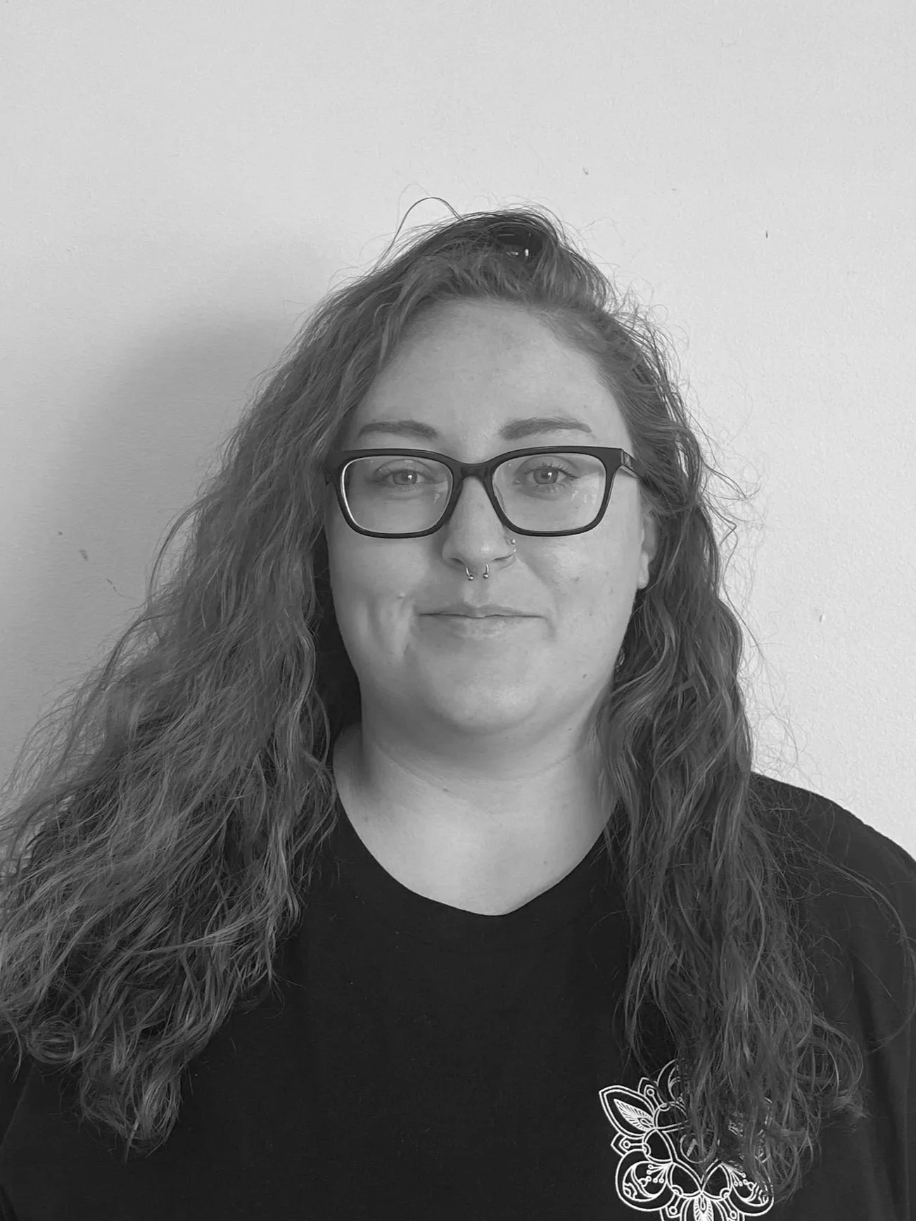 Black and white photo of a smiling young woman with long hair, wearing a black top, leopard print sleeves, and a cross necklace, standing against a plain wall.
