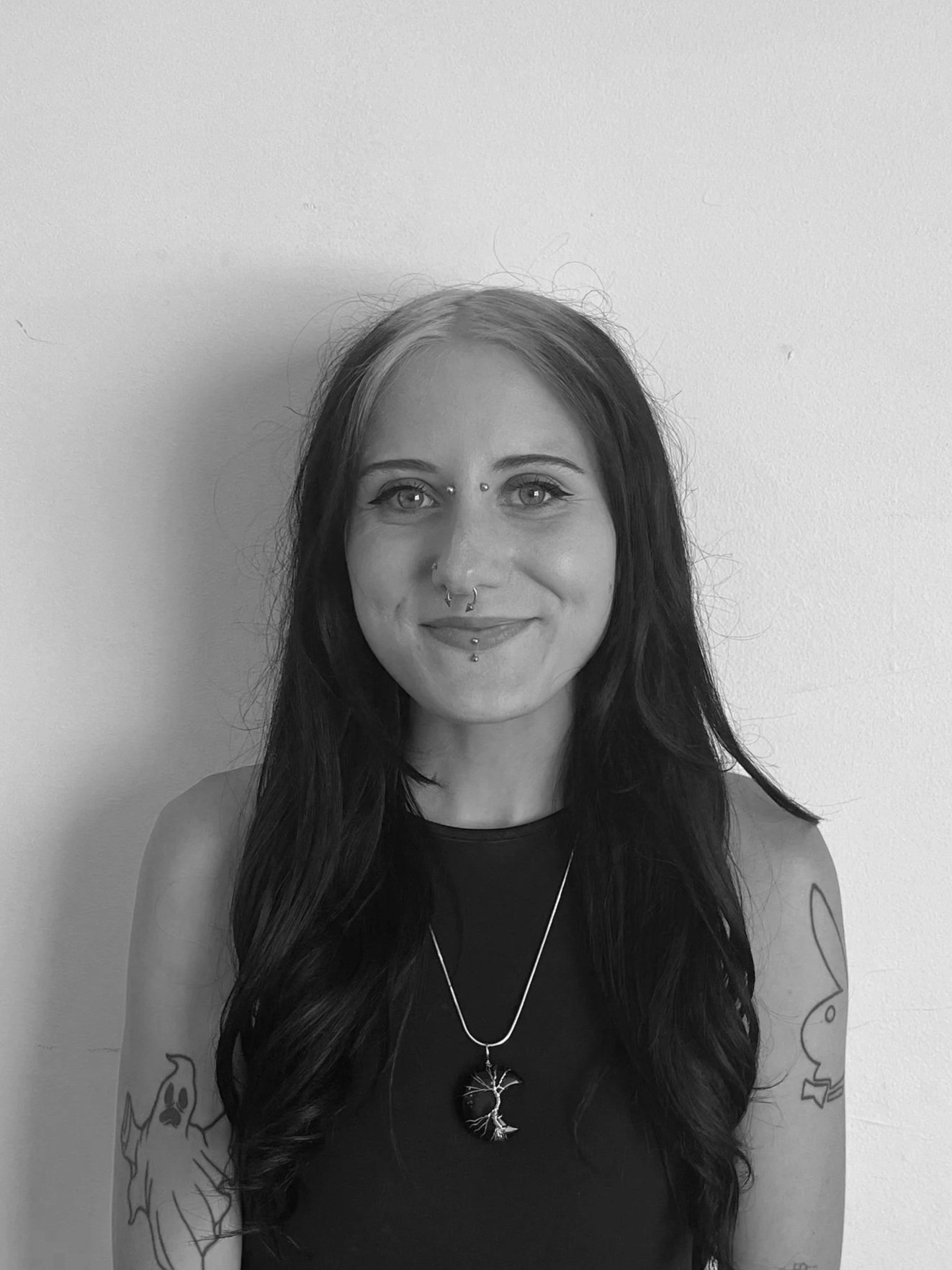 A young woman with long wavy hair, smiling, wearing a sweater, standing against a plain wall.