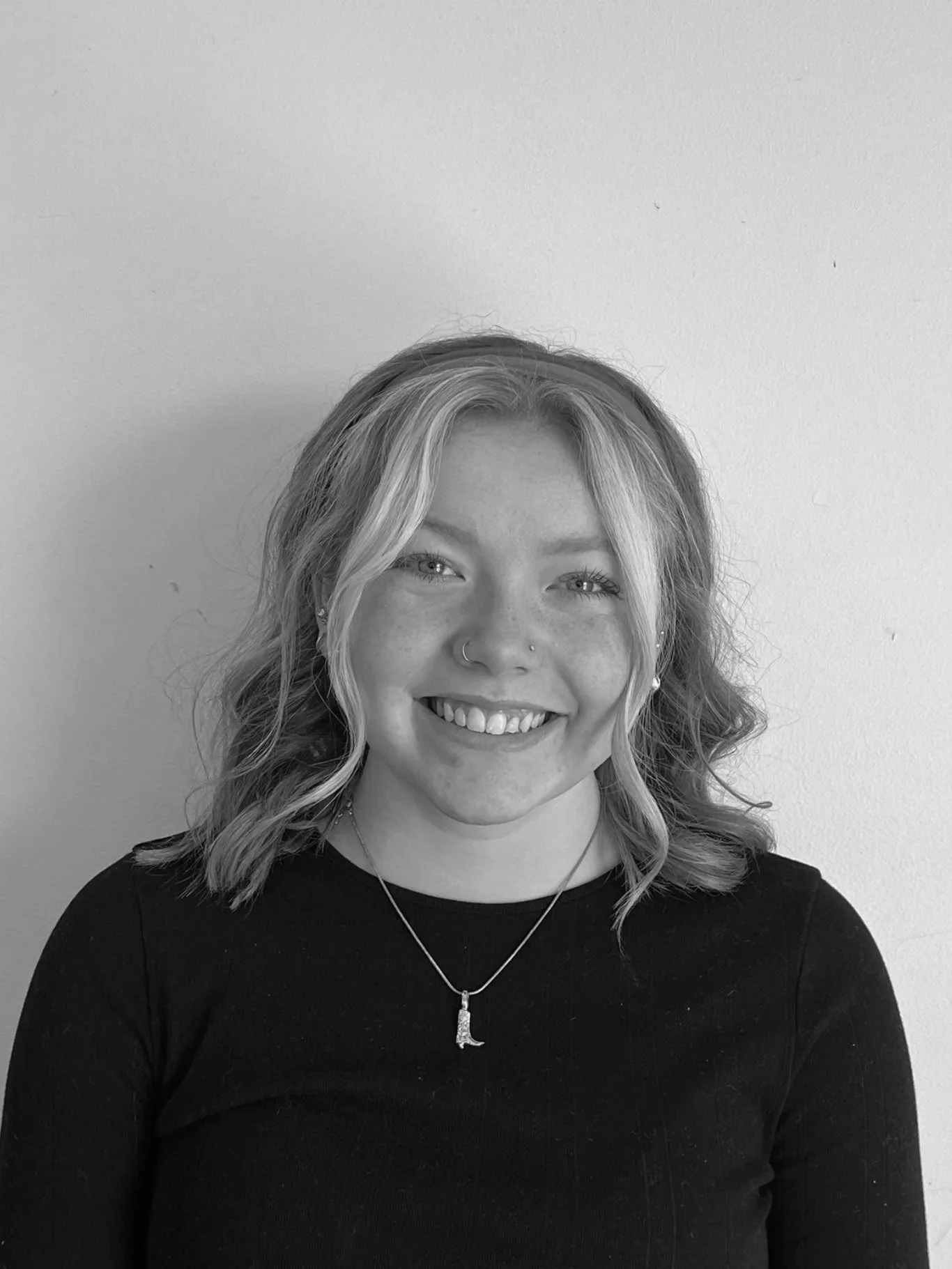 A young woman with glasses, long dark hair, wearing a black shirt, standing against a plain light-colored wall.