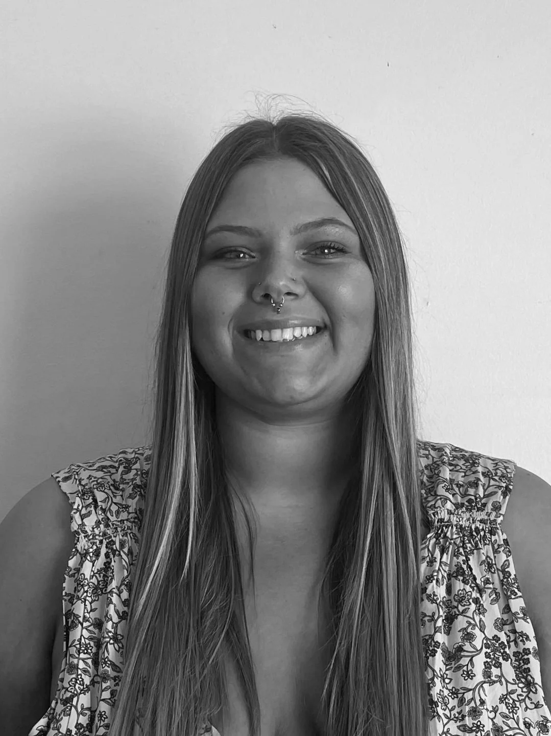 A black and white portrait of a young woman with long, straight hair and a nose piercing, smiling softly in front of a plain background.