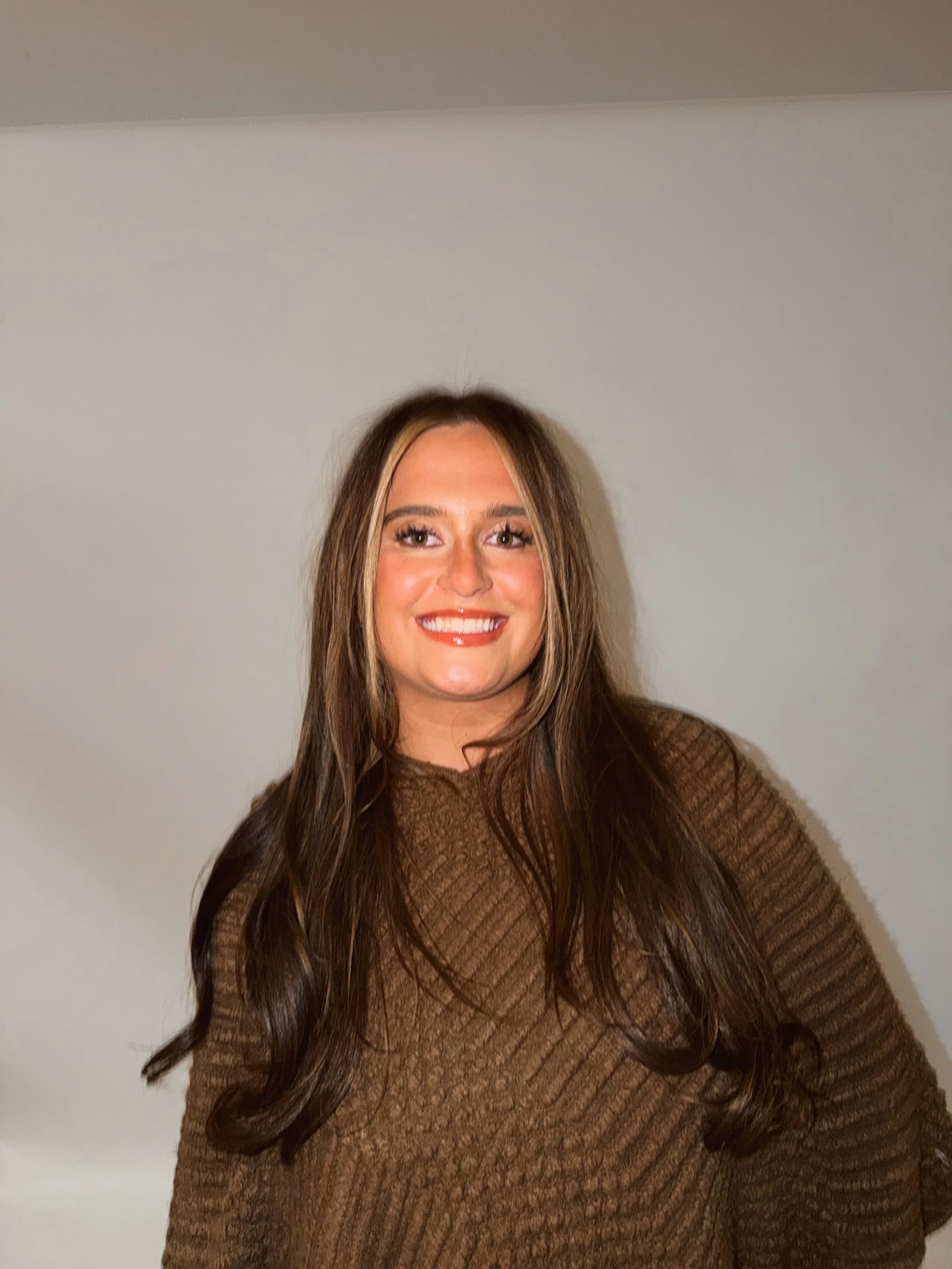 A woman with long wavy brown hair holding a Canon camera, standing outdoors near columns with a grassy area in the background.