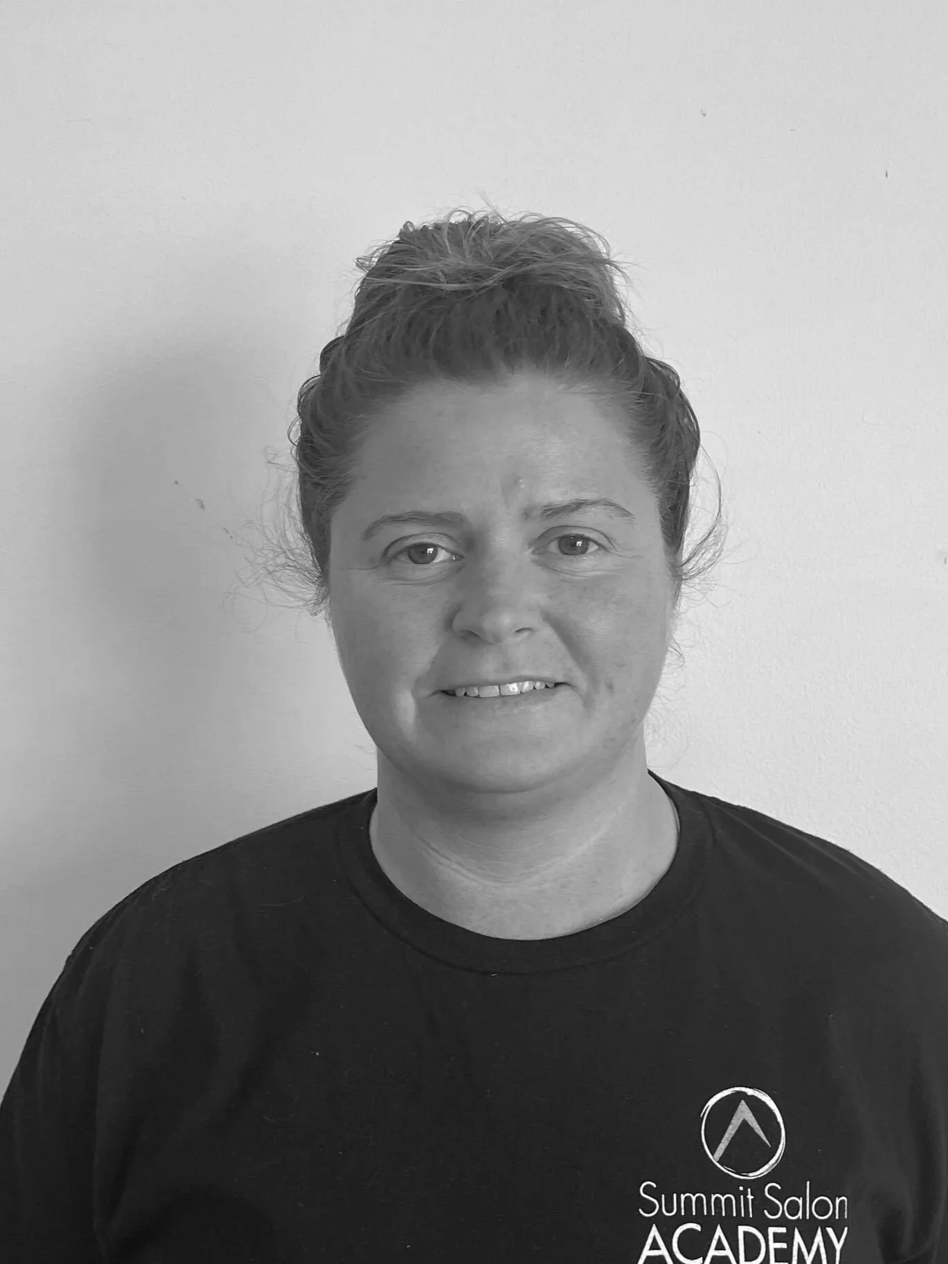 Black and white photo of a young woman with wavy hair, smiling, wearing a textured sweater, standing against a plain wall.