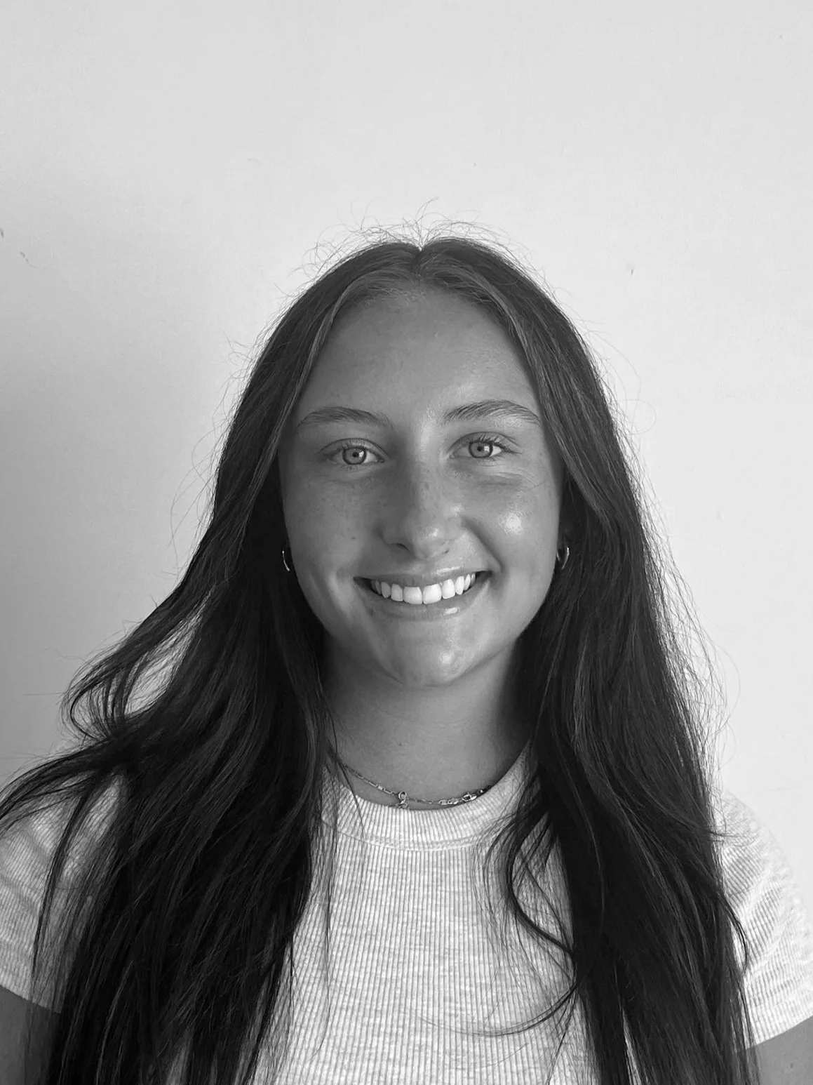 Black and white portrait of a young woman with long dark hair, smiling, wearing a denim jacket and a black shirt against a plain wall.