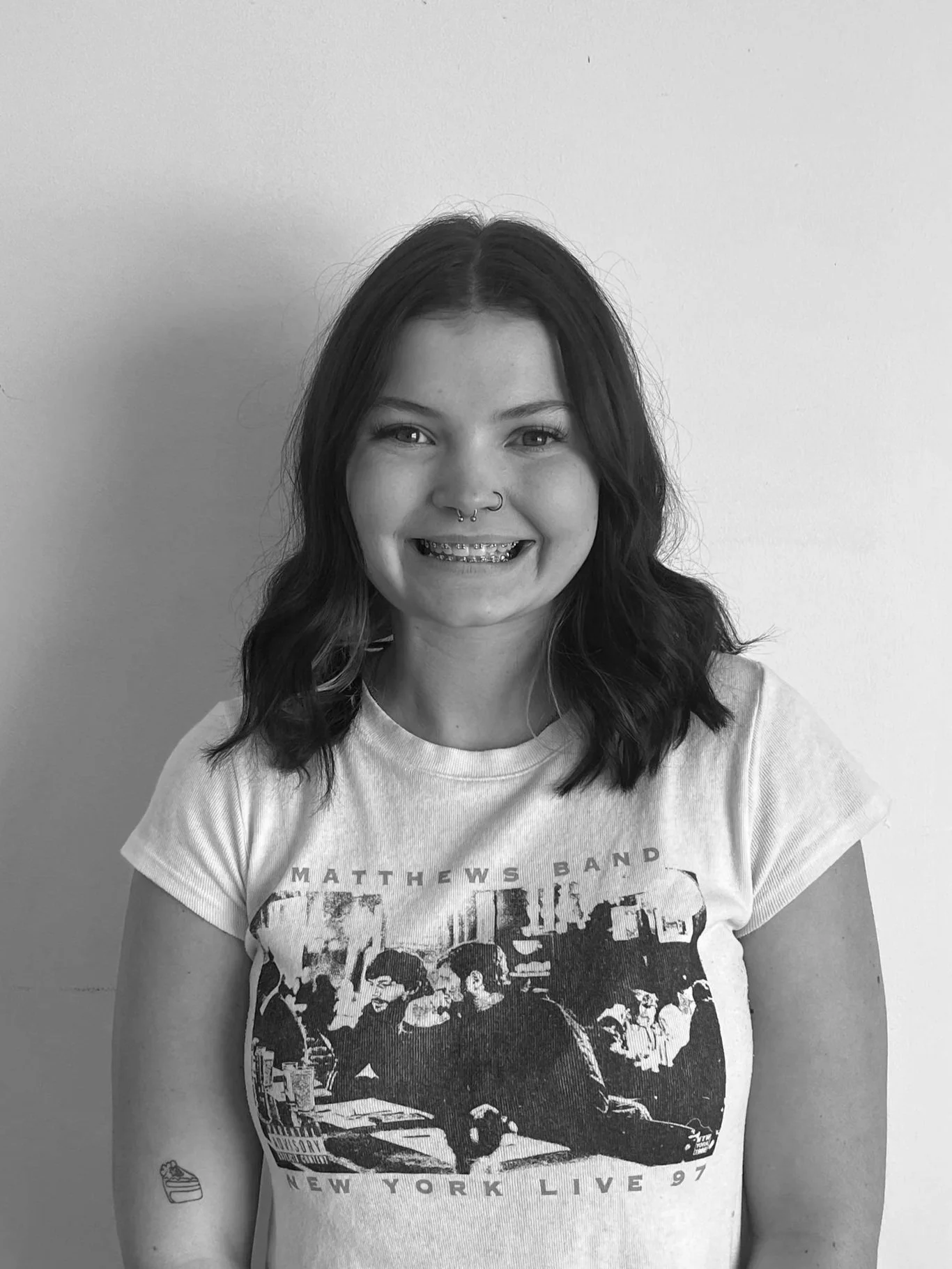 Black and white portrait of a smiling woman with shoulder-length wavy hair, wearing a plaid shirt over a black top, and layered necklaces, standing against a plain light-colored wall.