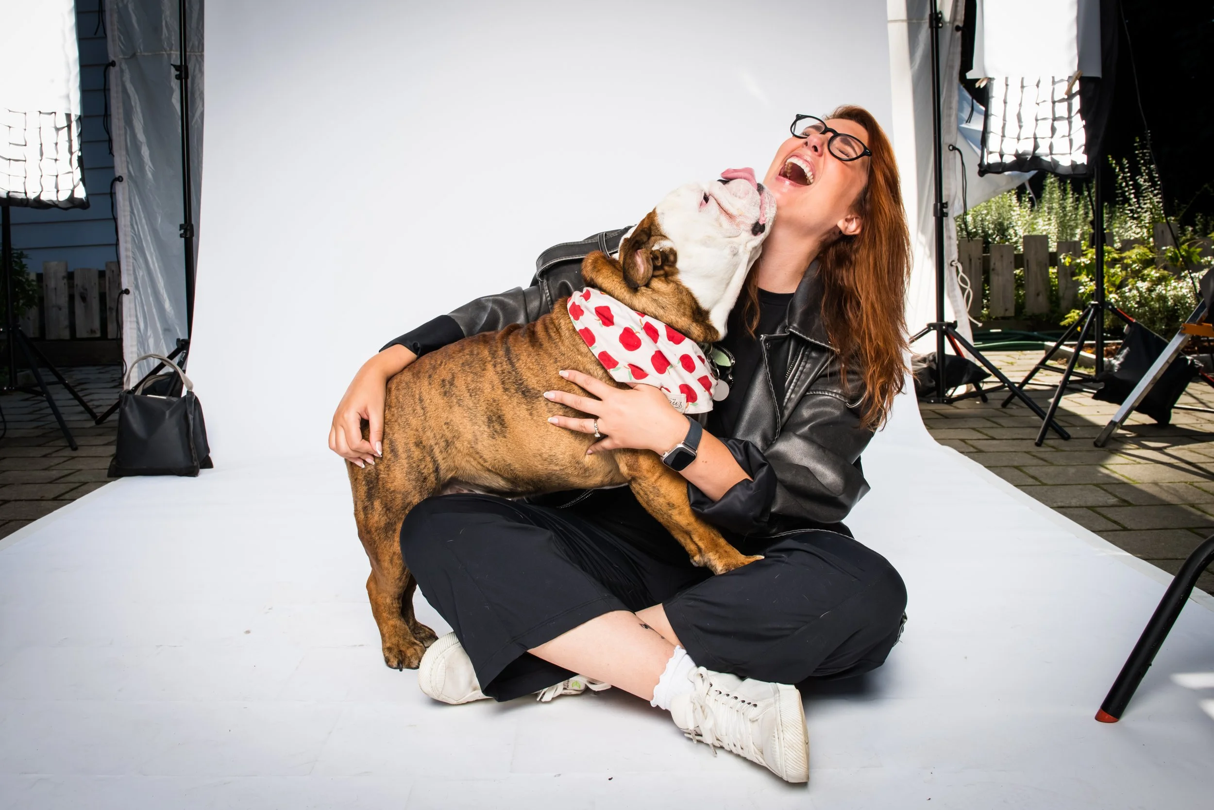 A woman with red hair and glasses is sitting on the floor, hugging a brindle bulldog that is licking her face. The dog is wearing a red and white heart-patterned bandana. The background shows professional photography equipment and a white backdrop, indicating a photo shoot is taking place.